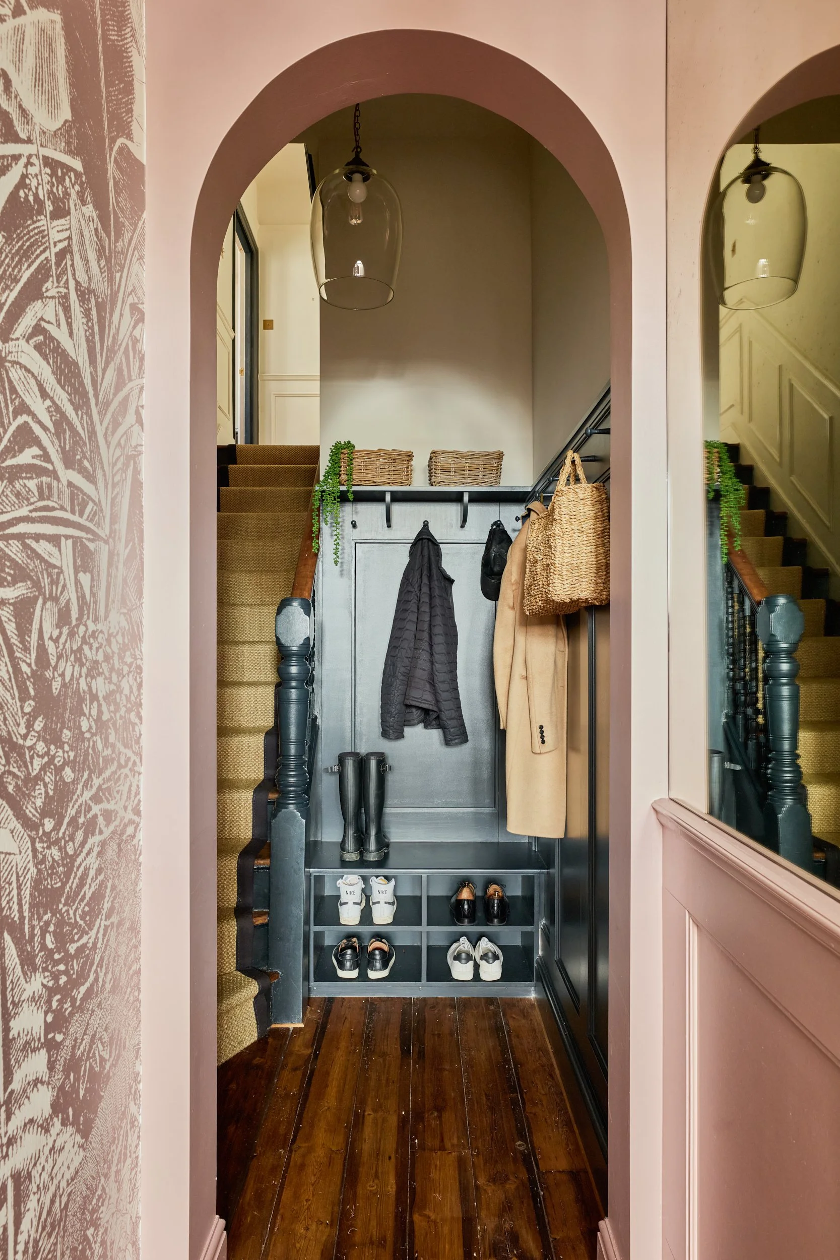 Entryway with stairs on both sides, black coat hooks on a gray cabinet, shoes in a cubby, hanging coats and bags, and pink walls.