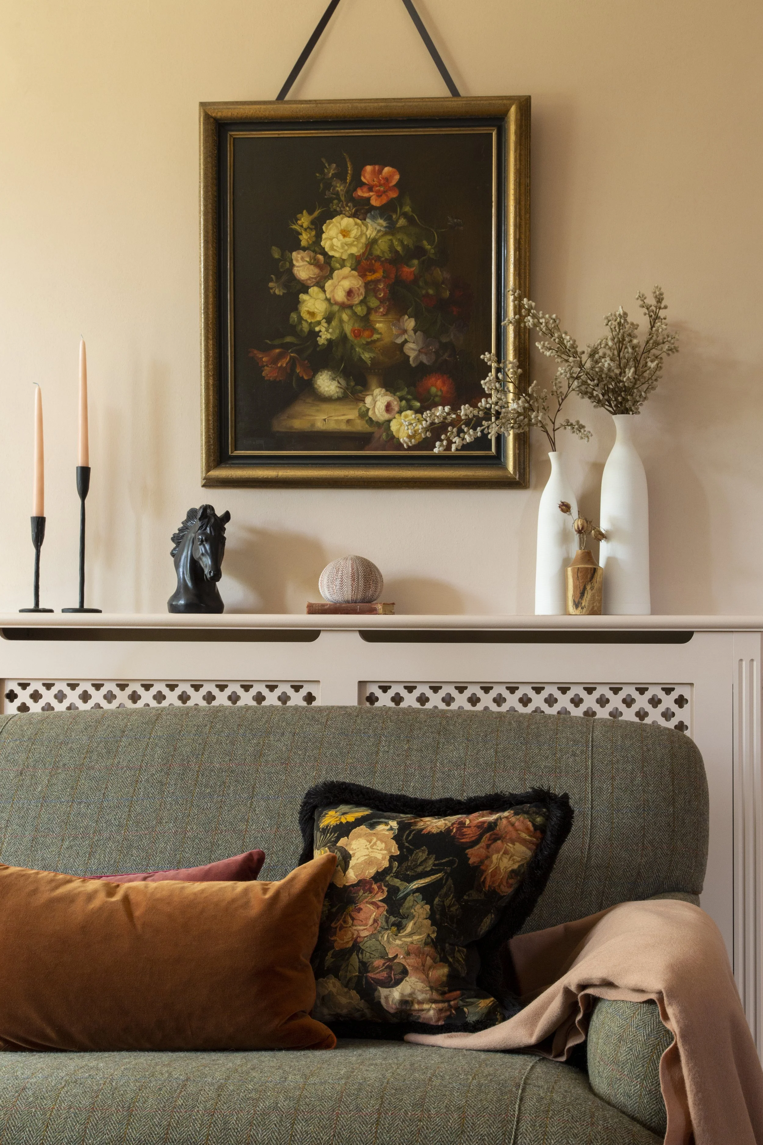 Living room corner with a gray couch adorned with a floral pillow, beige blanket, and brown pillow. A white mantel behind the couch holds decorative items: black horse head sculpture, a round textured ball, two white vases with dried flowers, and two pink candles. A framed floral still life painting hangs above the mantel.