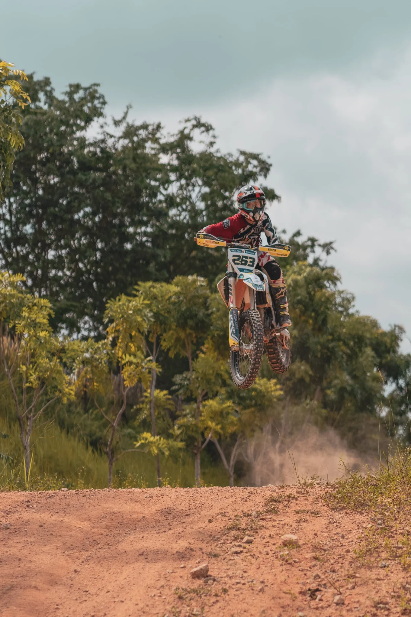 Motocross rider in red gear and helmet jumps off a dirt track with trees and a cloudy sky in the background.