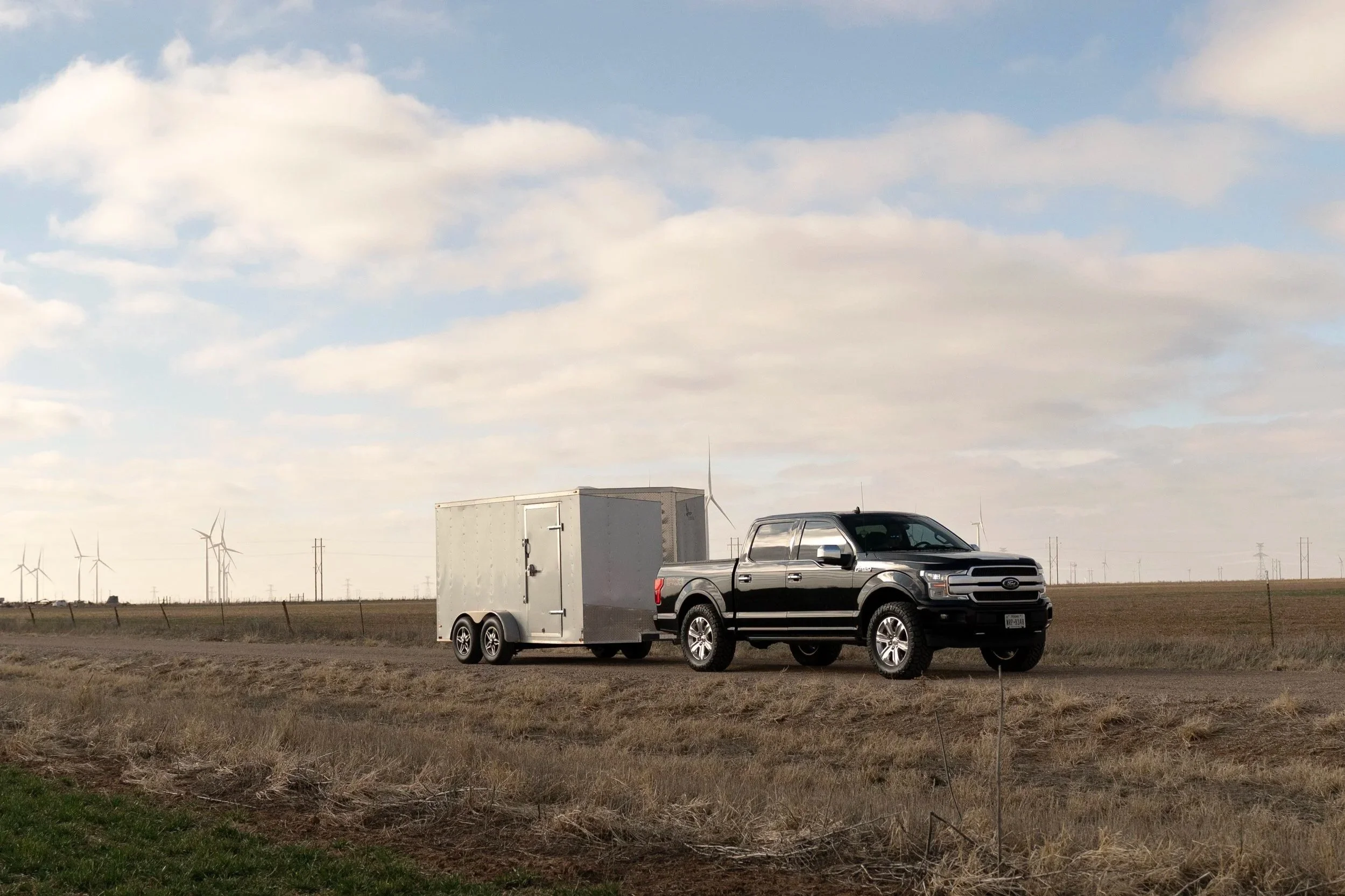 A black pickup truck attached to a silver enclosed trailer is parked in an open field. In the background, there are wind turbines and a partly cloudy sky.