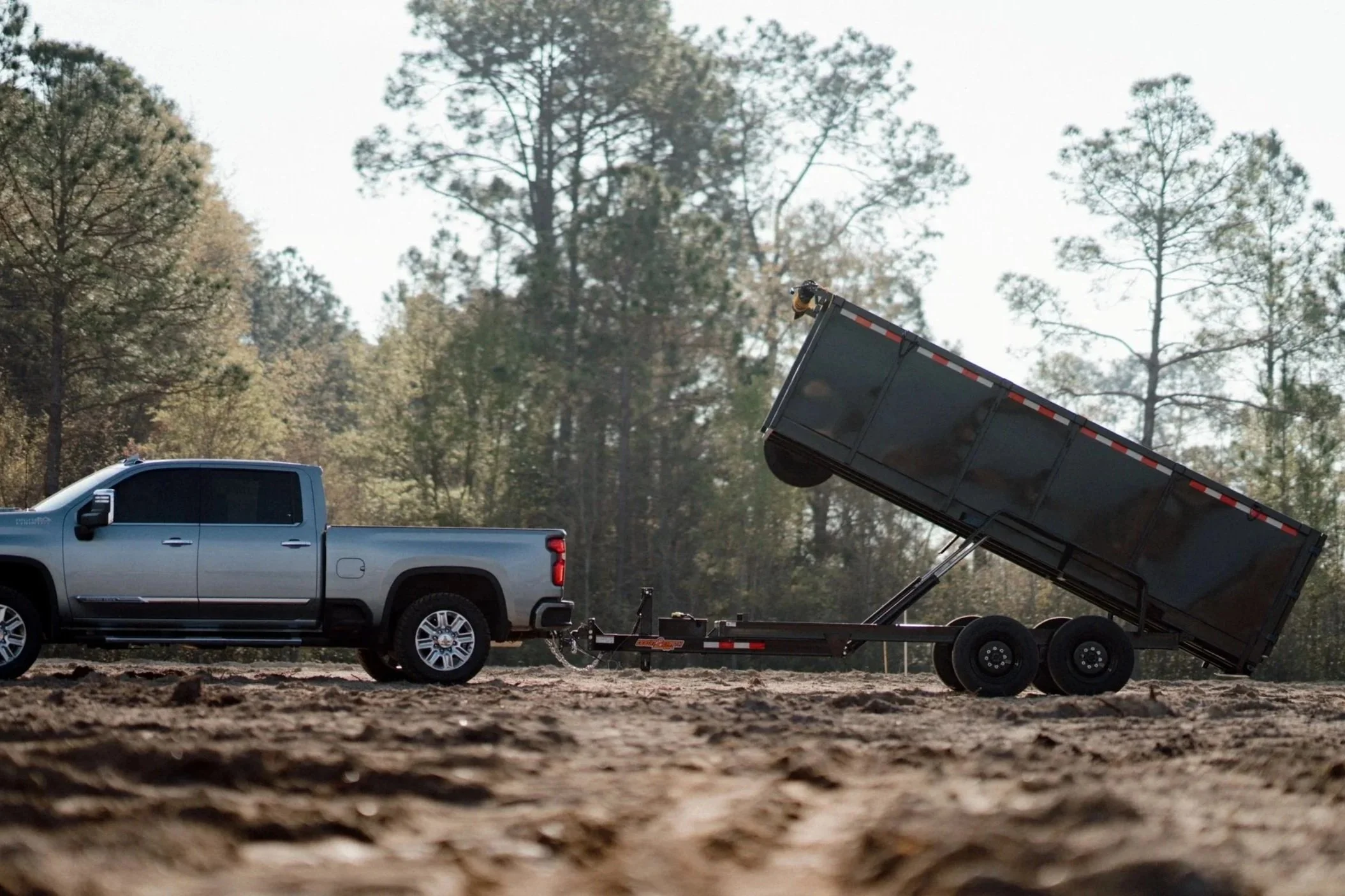 A silver pickup truck pulling a flatbed trailer with a raised dumping bed in a wooded outdoor area.