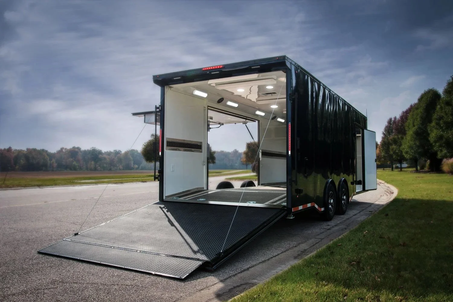 A black enclosed trailer with a fold-down ramp parked on a paved road, open to show the interior with lighting and two wheels stored inside.
