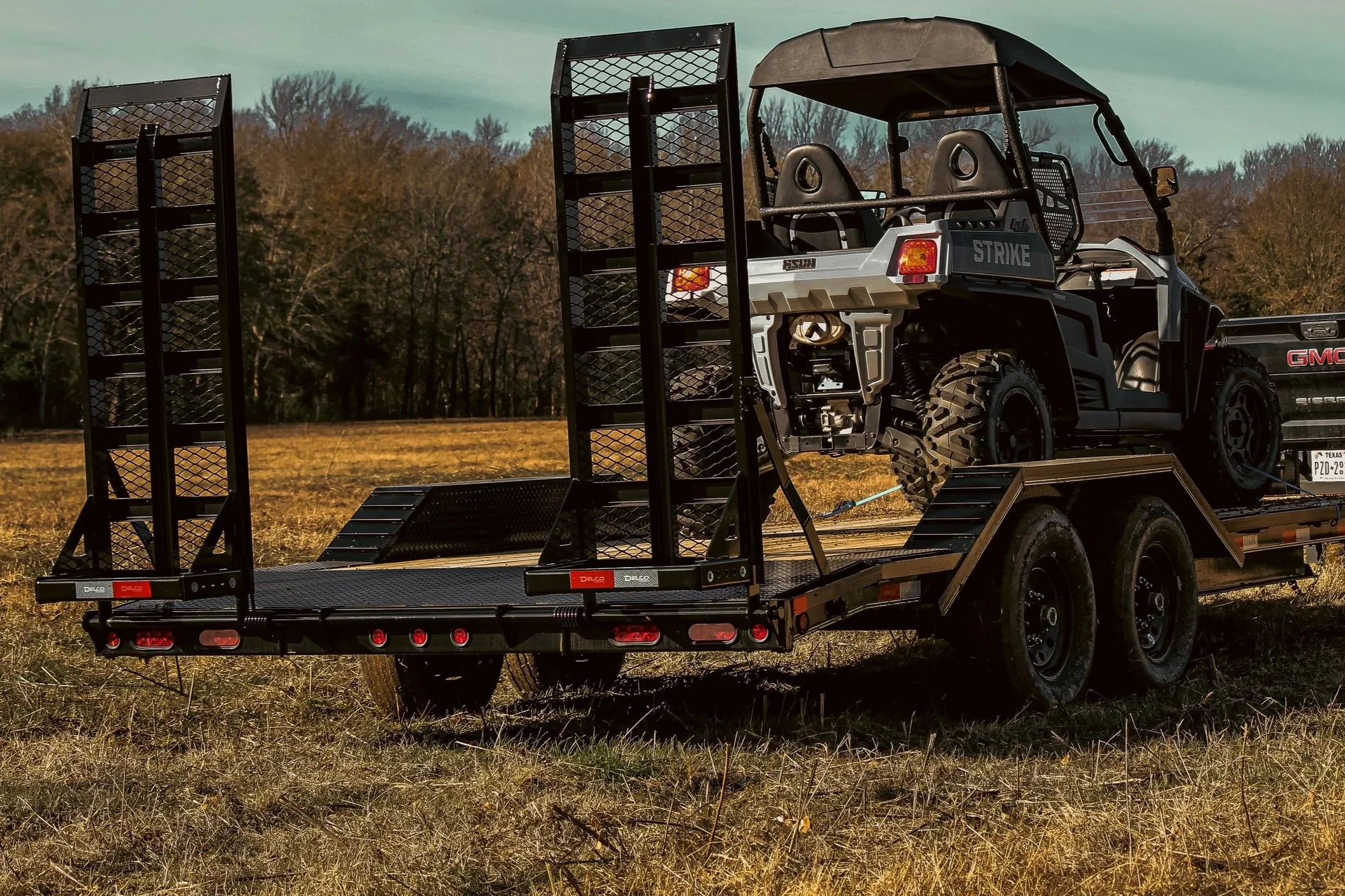 A black GMC Utility Task Vehicle (UTV) loaded on a flatbed trailer with ramps, parked in an open field with trees in the background on a clear day.