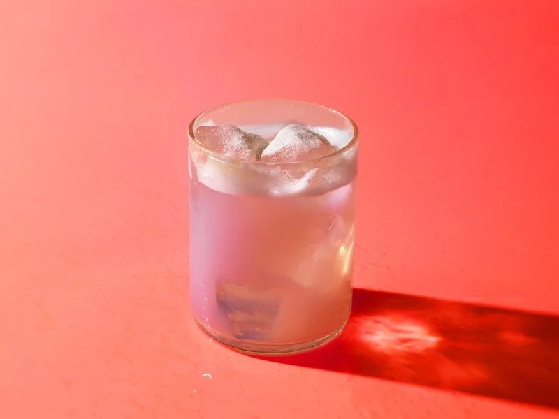 Pink ARMRA colostrum drink with ice in glass on coral background