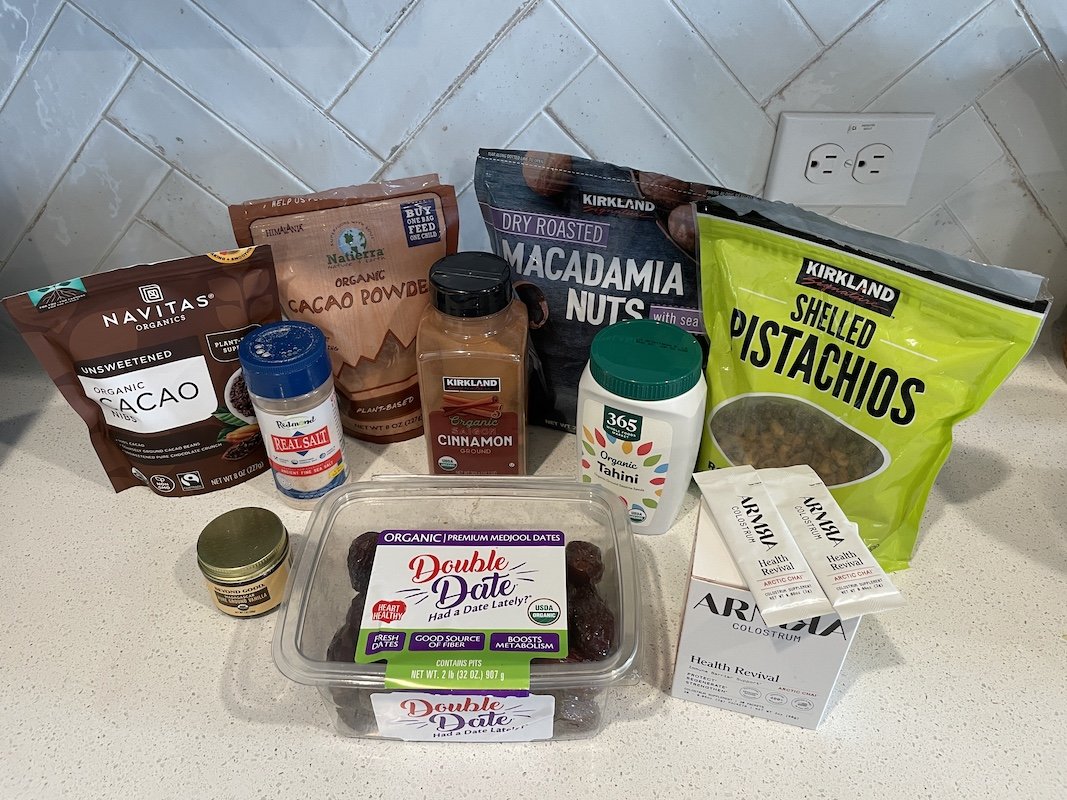 Ingredients for no bake Armra colostrum date bars laid out on a kitchen counter, including dates, tahini, cacao, macadamia nuts, and pistachios