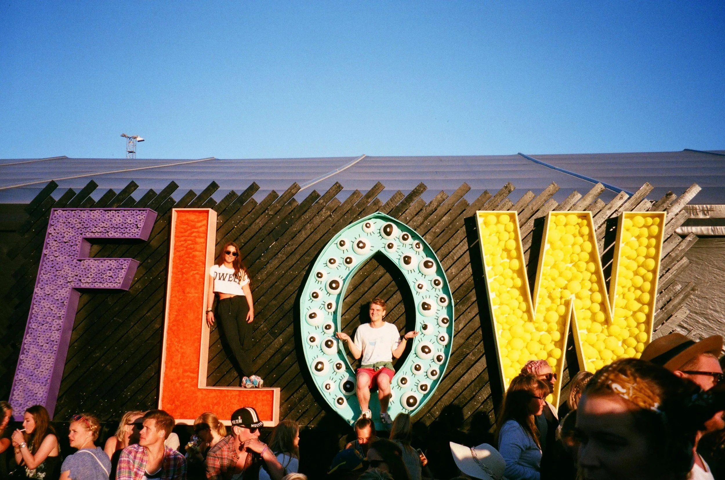Giant letters spelling FLOW at an outdoor festival, people posing underneath