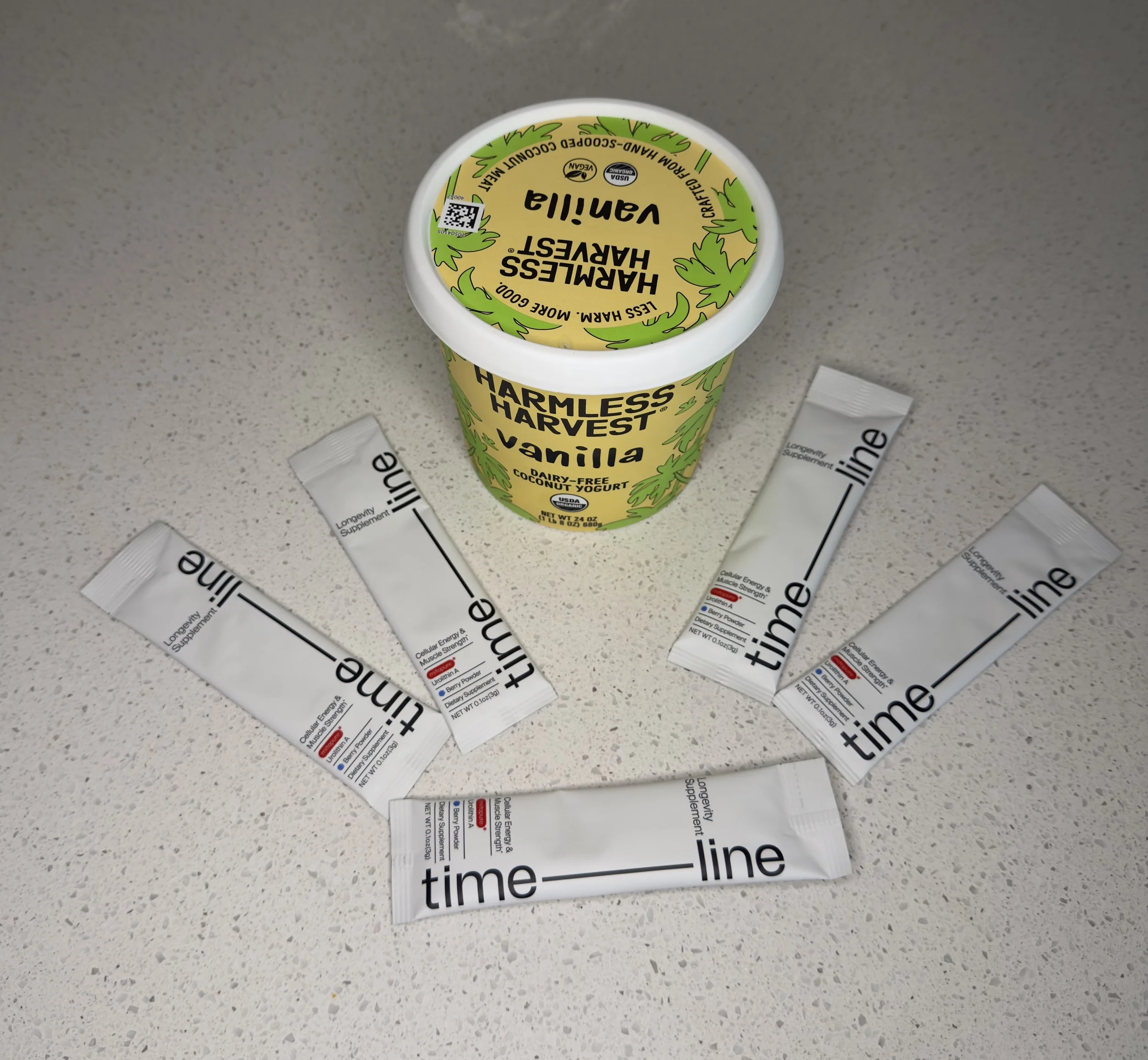 Timeline Mitopure powder sachets fanned out next to a tub of Harmless Harvest vanilla coconut yogurt
