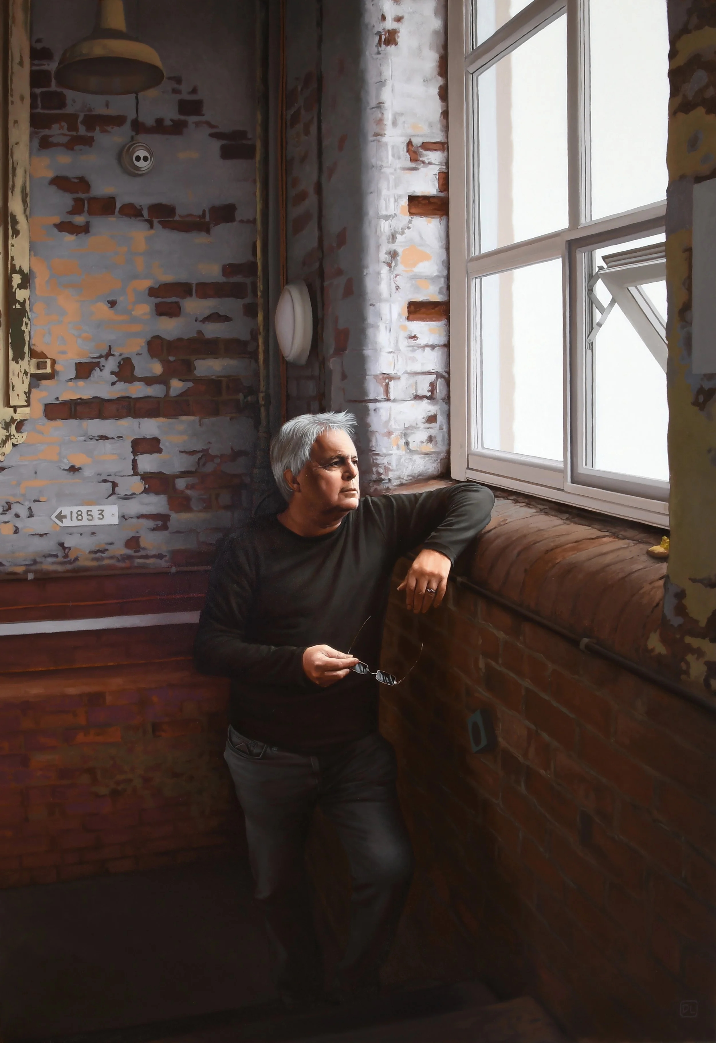 A man with gray hair, wearing a black long-sleeve shirt and gray pants, standing next to a window in a brick-walled room, looking outside with a contemplative expression, holding glasses in his right hand.