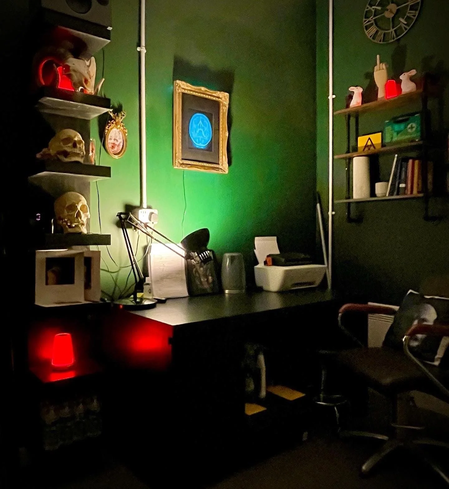 A dimly lit office or workspace with dark green walls. On the left wall, there are shelves holding skull models, a mask, and various decorative items. The desk has a lamp, keyboard, printer, and other office supplies. There is a framed digital artwork or logo on the wall. Red decorative lights are visible on the bottom shelves. A clock is mounted on the upper right wall.