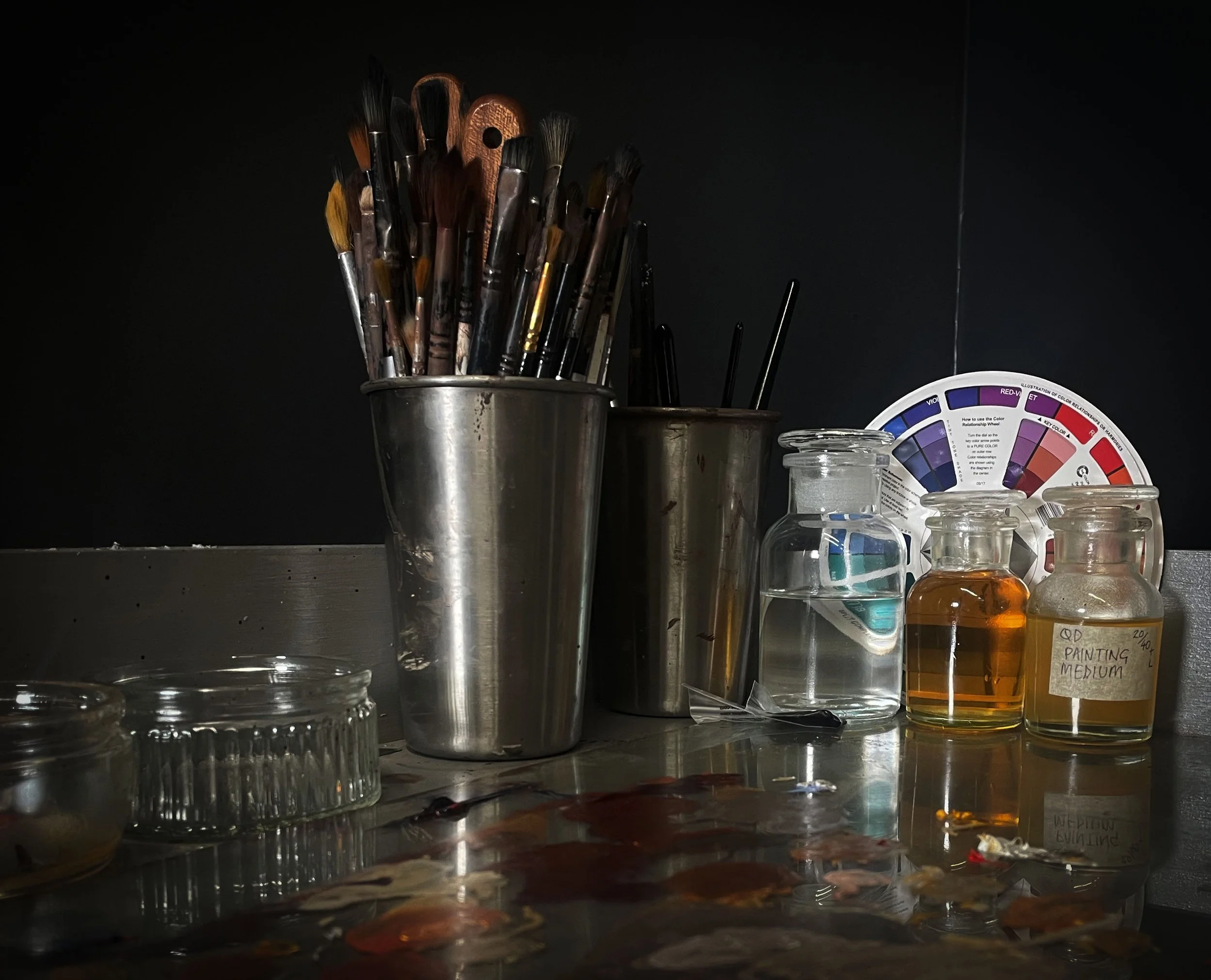 Artist's work station with paintbrushes in metal containers, bottles of water and paint medium, and a color wheel.