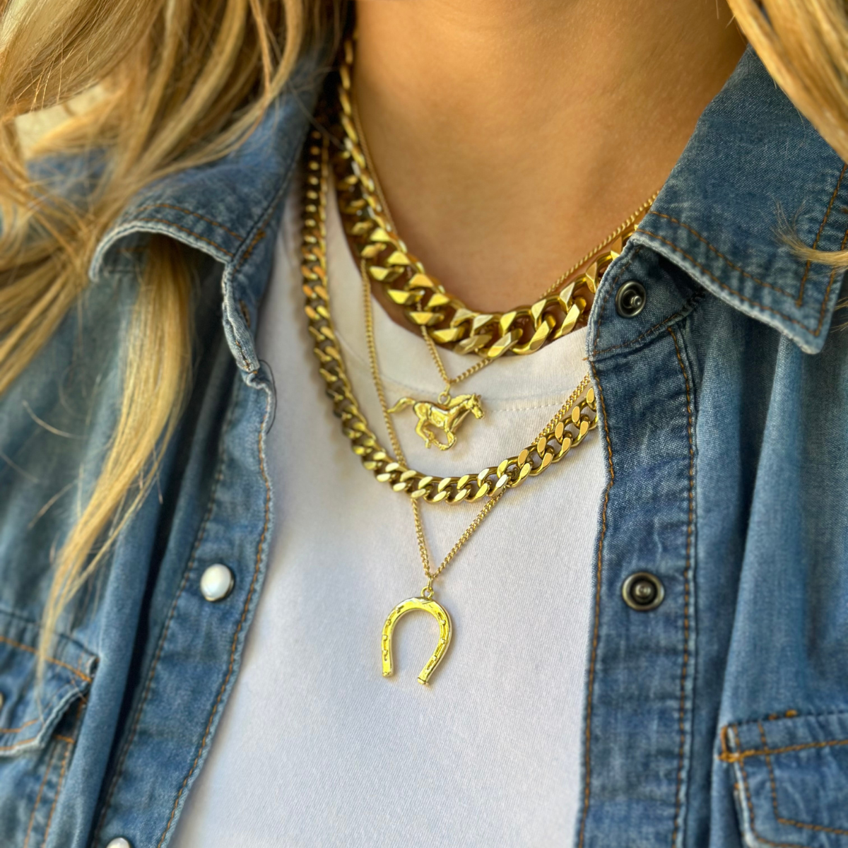A person wearing layered gold necklaces, including a chunky chain, a horse pendant chain, a horseshoe necklace, and a small chain. They are dressed in a white top and a denim jacket.