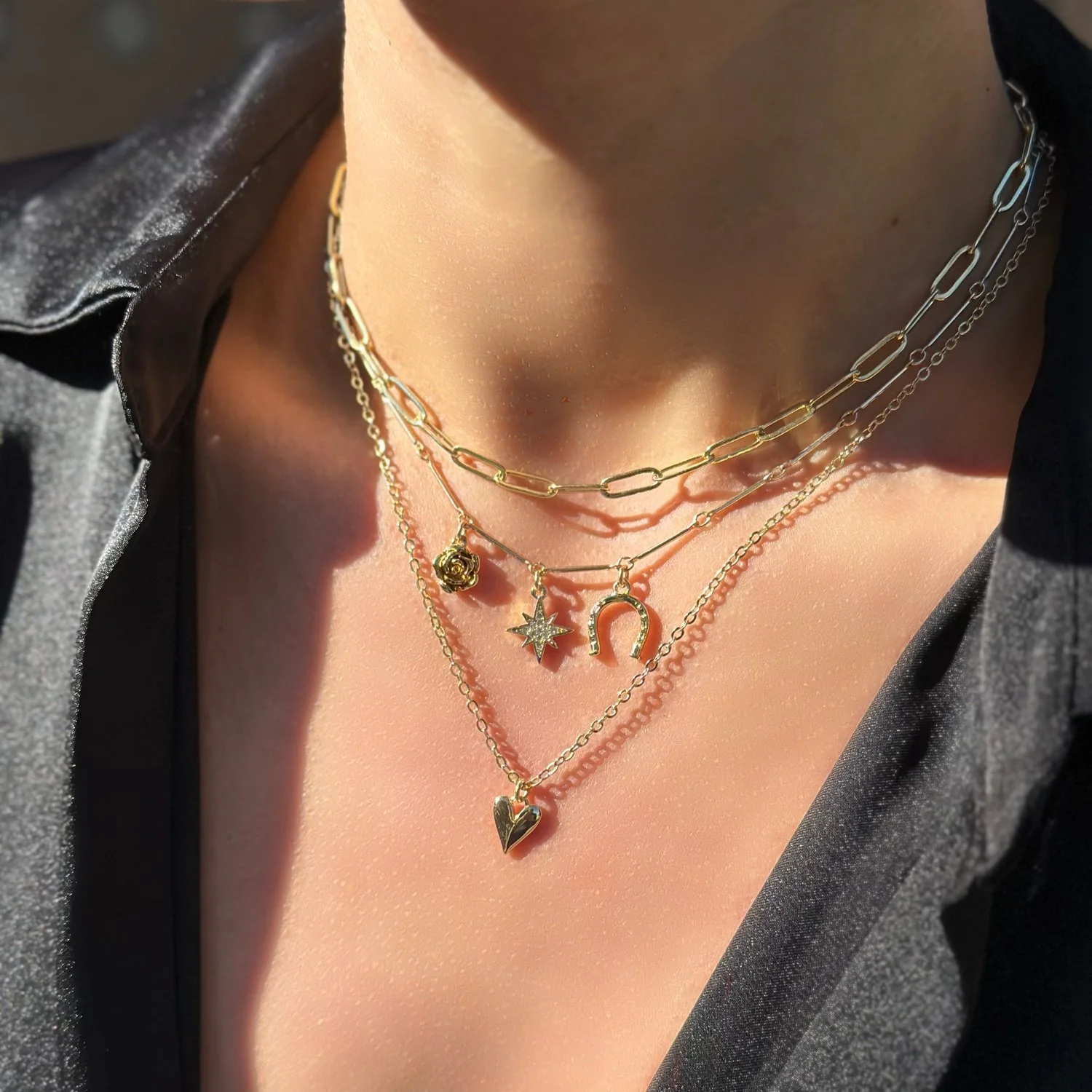 Multiple layered gold necklaces with various charms, including a rose, star, horseshoe, heart, and a dangling star, worn over a person's neck and chest.