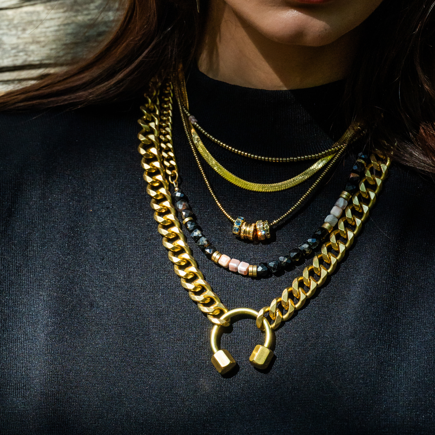Close-up of woman's neck and chest showing layered gold and beaded necklaces against a black fabric background.
