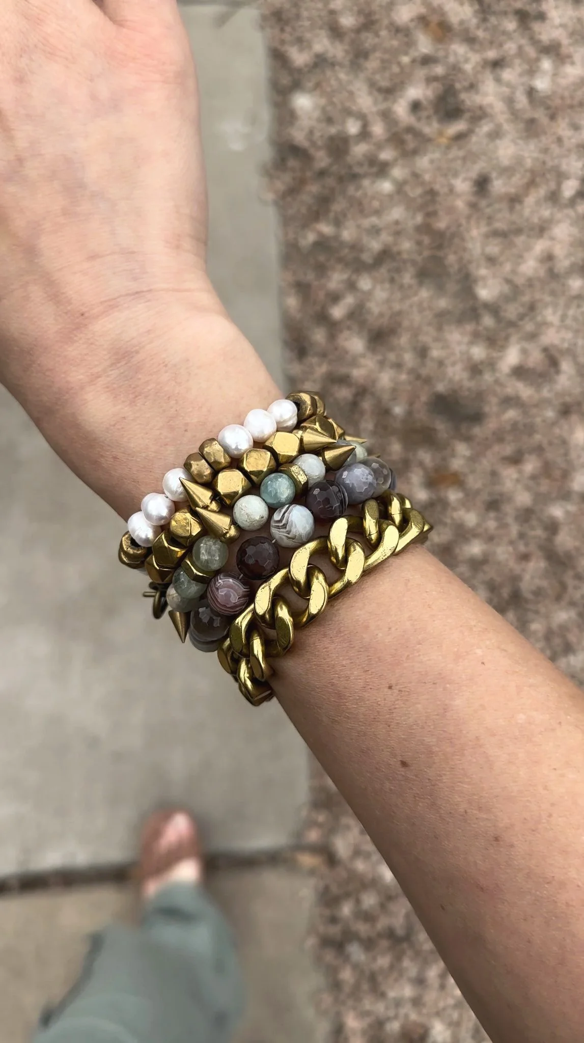 A person’s wrist with four stacked bracelets: a gold chain link bracelet, a bracelet with gold and black beads, a beaded bracelet with light green, gray, and white beads, and a bracelet with white pearls. The background is a concrete sidewalk and a stone wall.