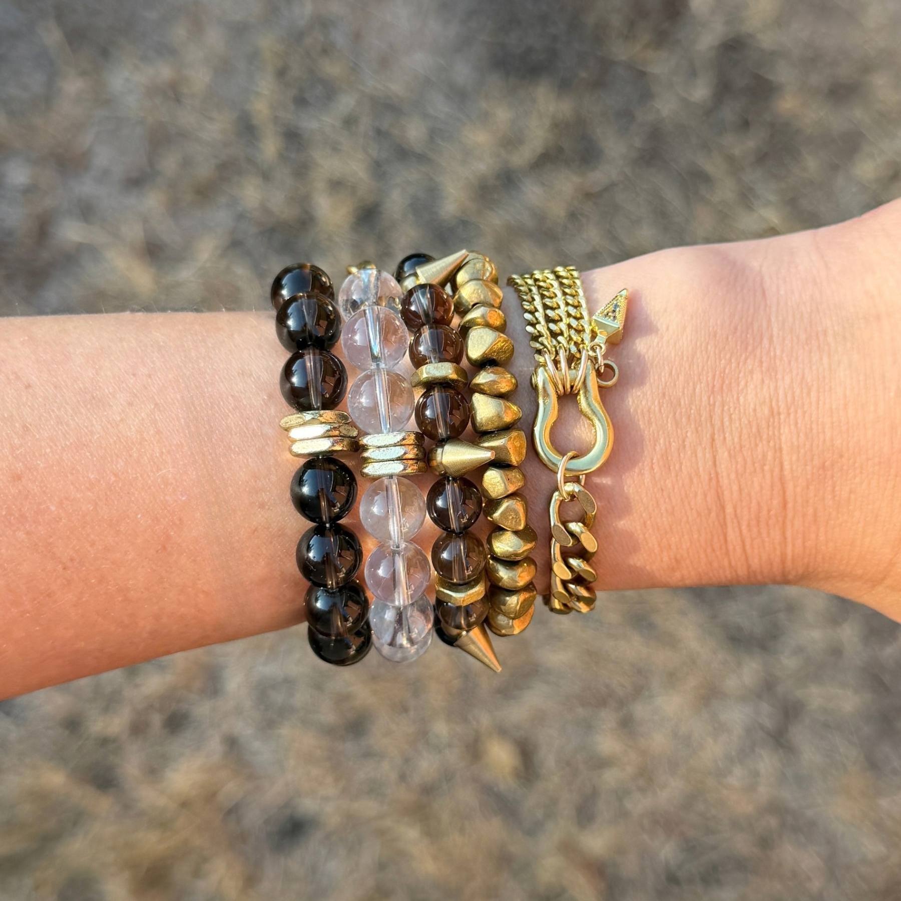 A person's wrist wearing multiple bracelets, including black beads, pink beads, gold beads, and a gold chain bracelet with a hook.