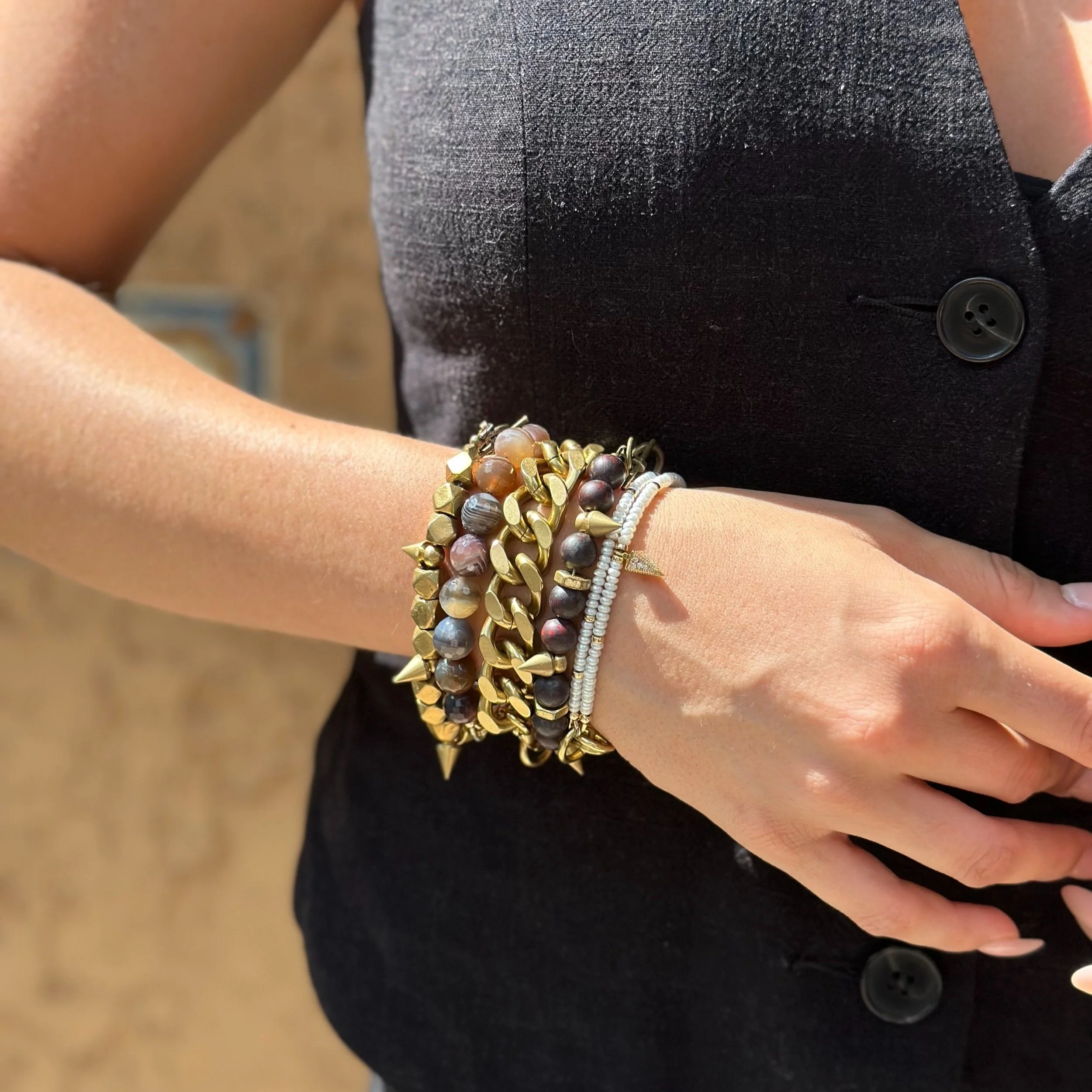 Close-up of a person's wrist adorned with multiple bracelets, wearing a black dress or jacket with buttons, against a blurred outdoor background.
