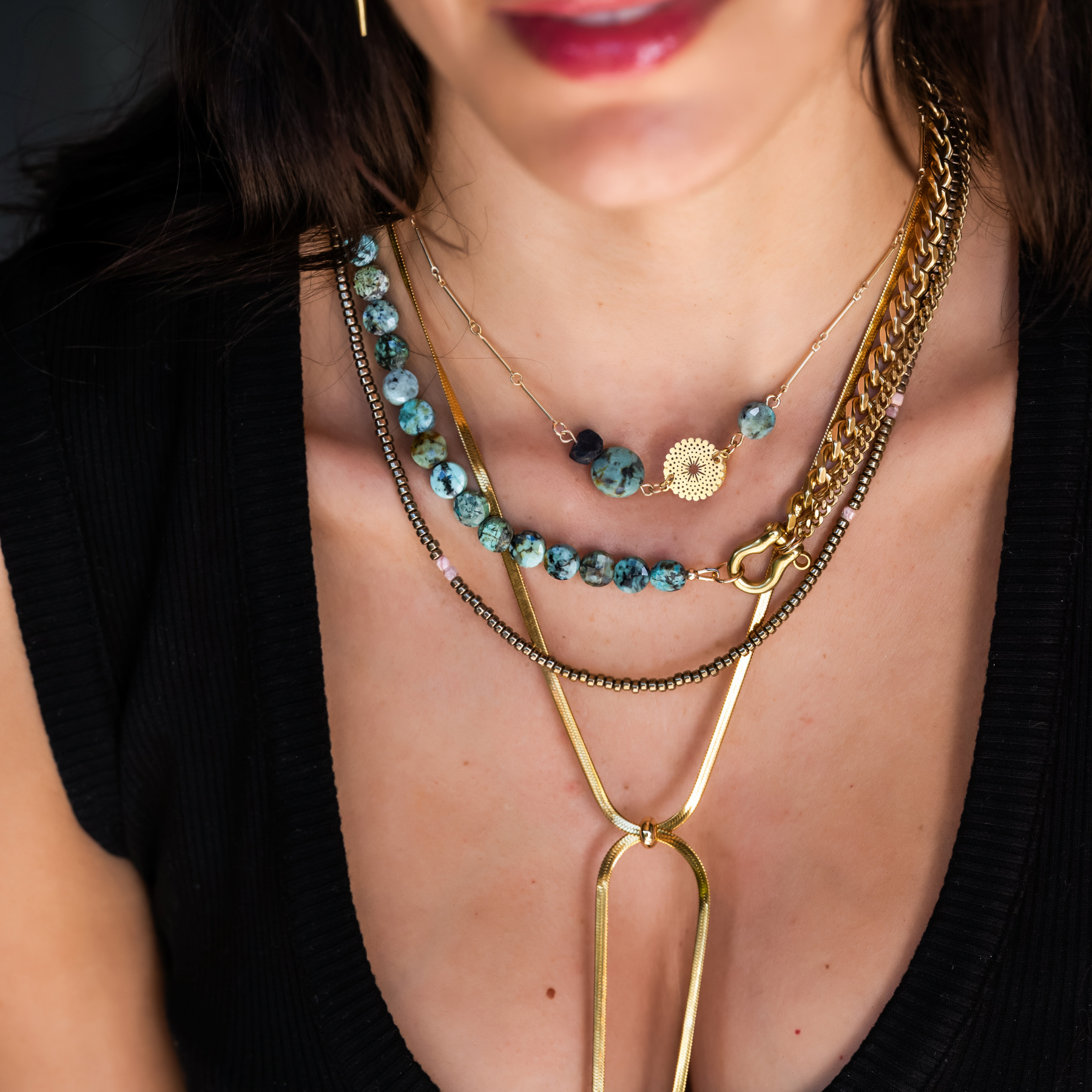 Close-up of a woman's neck and chest area, showing layered necklaces including turquoise beads, gold chains, and a circular gold pendant, with part of her face visible including lips and hair.
