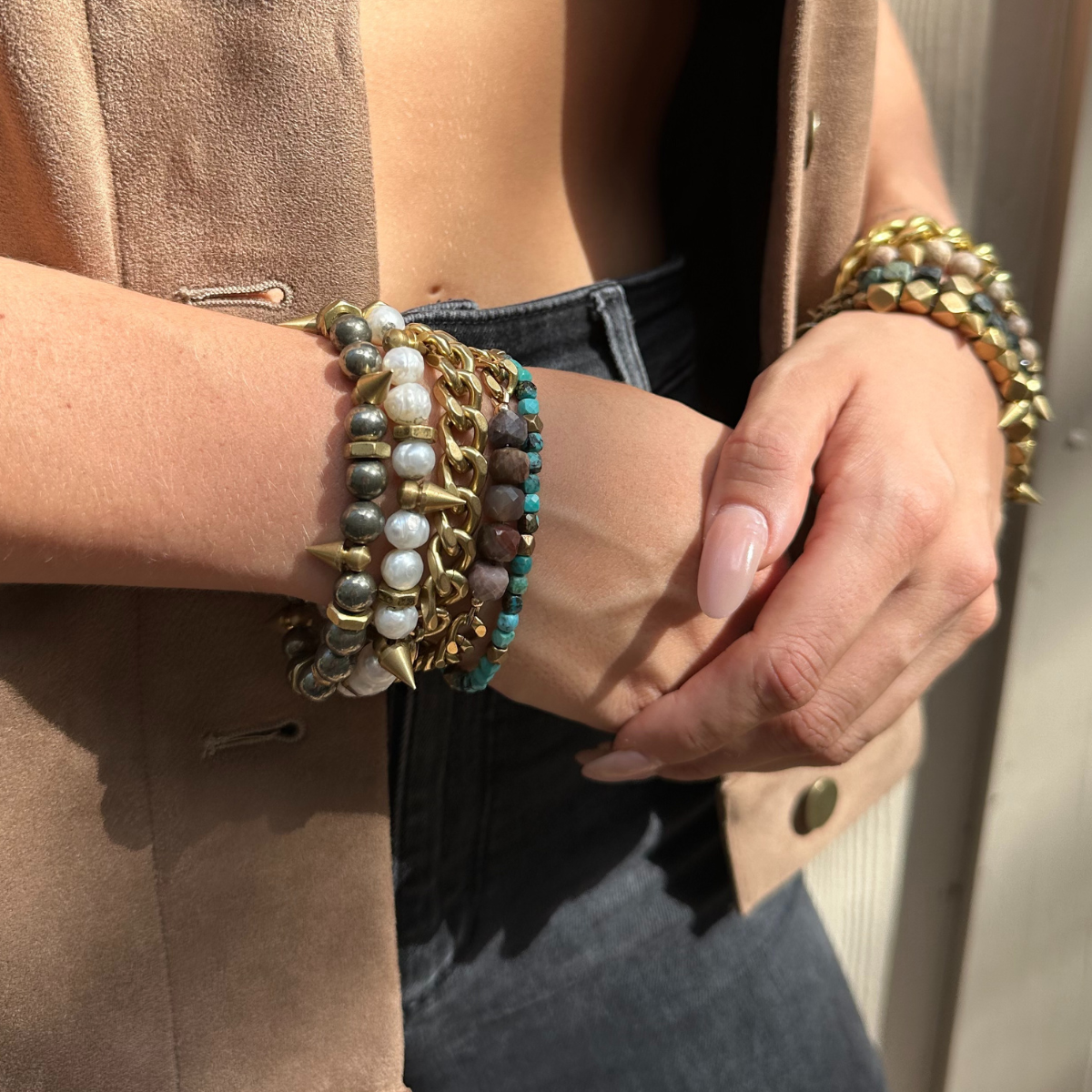 Person wearing multiple bracelets on left wrist, including beads, pearls, and studs, with hand resting on stomach. Person is dressed in a tan jacket, black top, and dark jeans.
