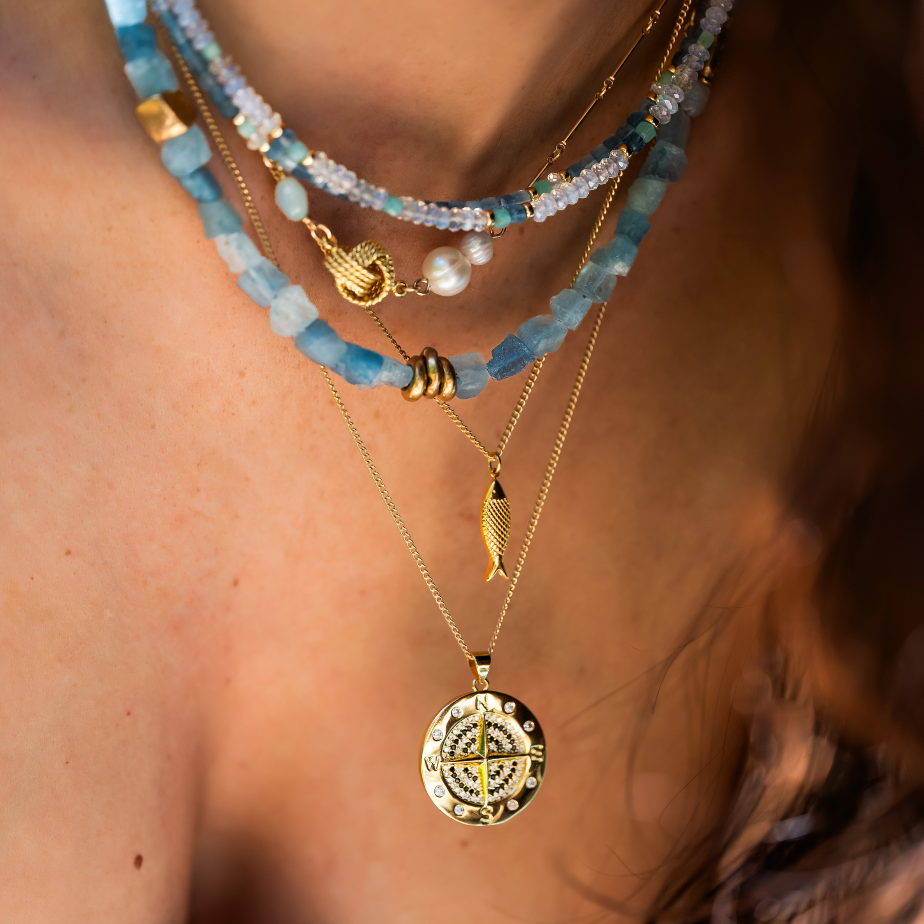 Close-up of layered gold and beaded necklaces on a person's neck, featuring various charms including pearls, a peace sign, a fish, and a circular design with stones.