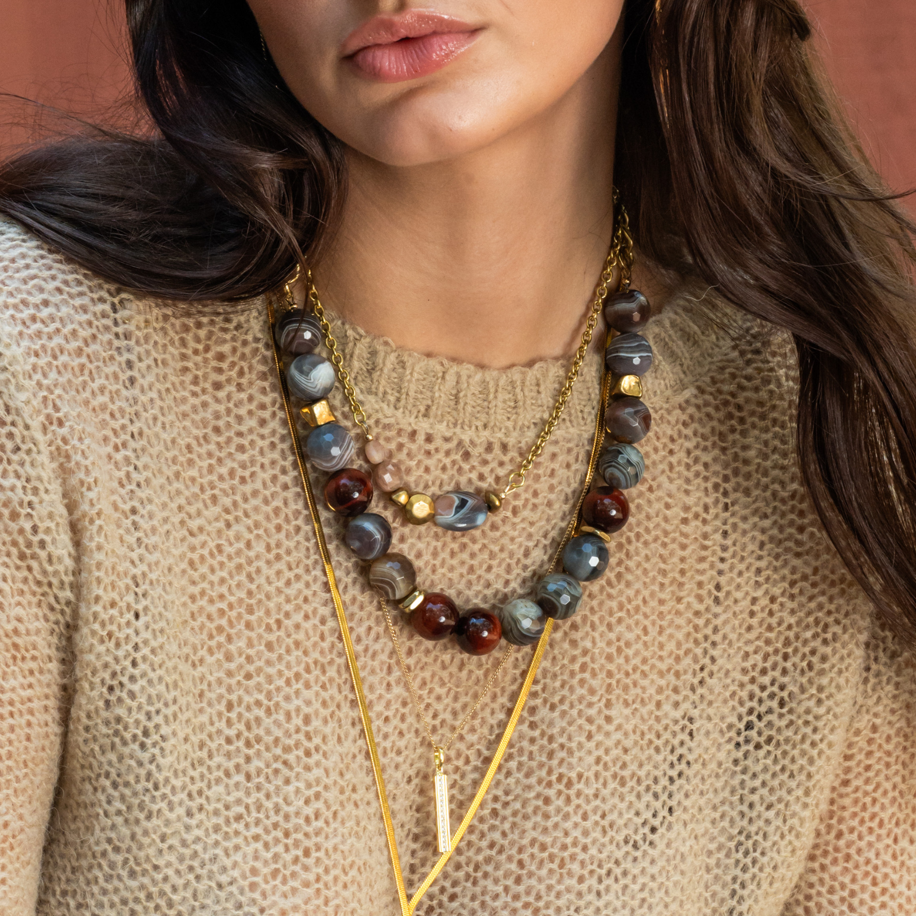 A close-up of a woman's lips, chin, and necklaces. She is wearing a beige knitted sweater and layered gold necklaces with large, dark, patterned beads.