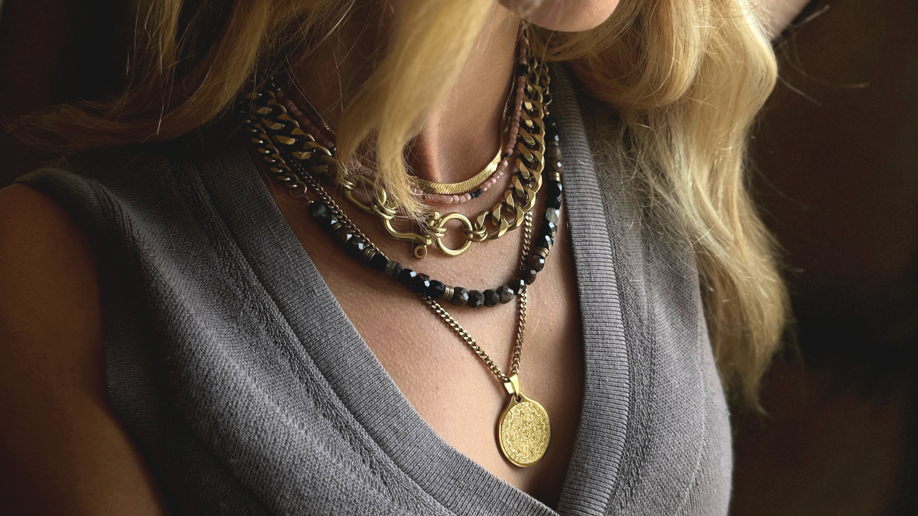 Close-up of a woman's neck and upper chest wearing multiple layered necklaces, including gold chains, beaded necklaces, and a pendant with intricate design, over a grey top.