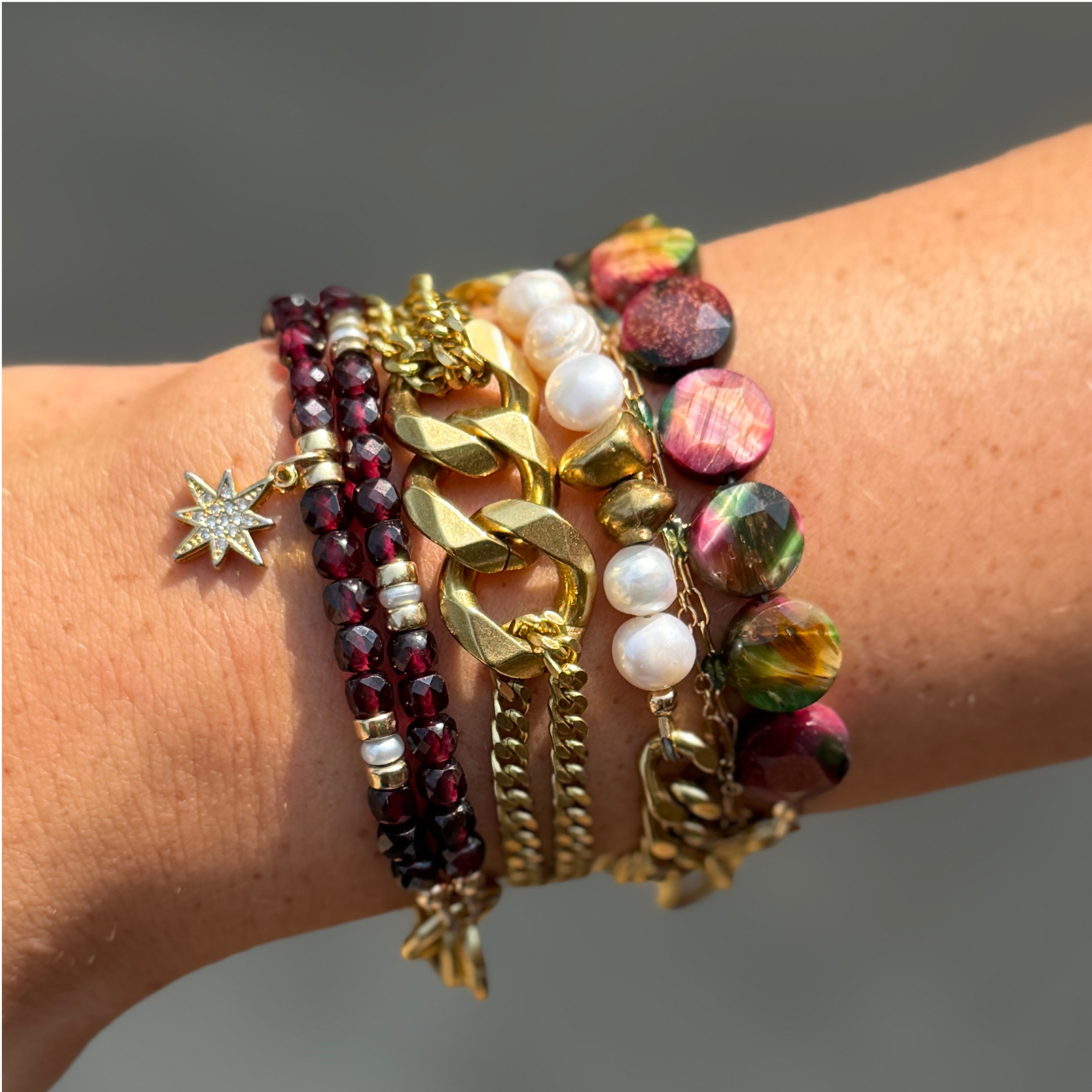 Multiple bracelets on a person's wrist, including a gold chain bracelet, a beaded bracelet with white pearls, a bracelet with dark red beads, and a colorful bracelet with pink and green stones.