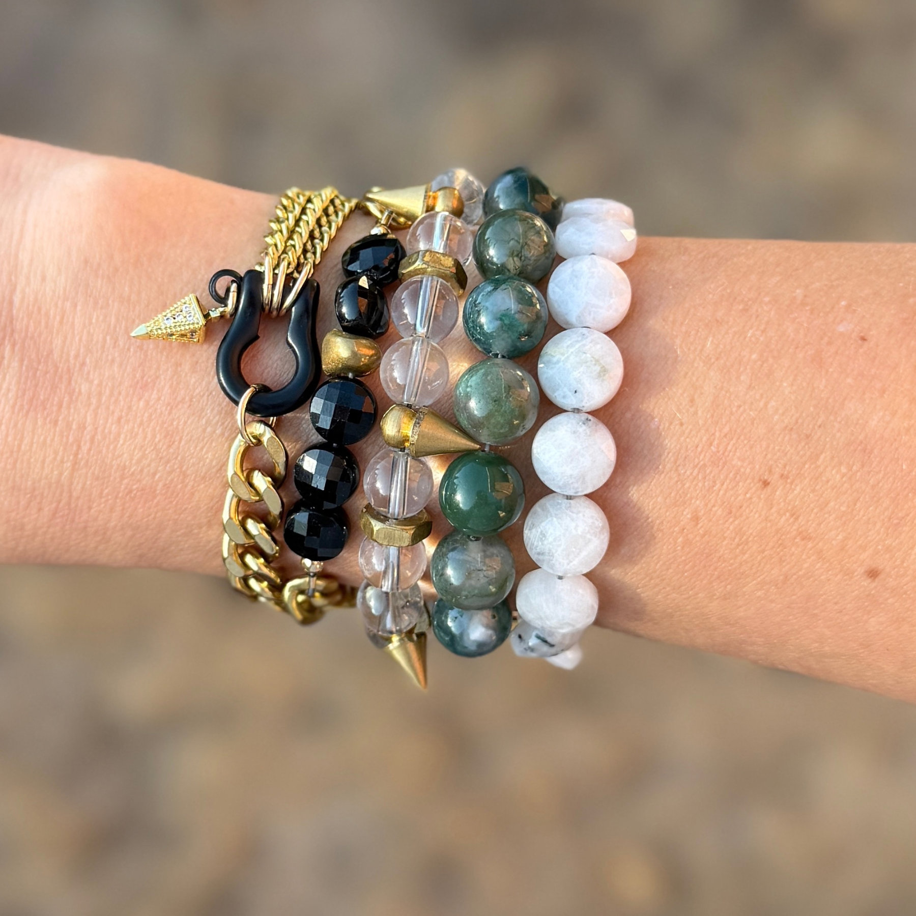 Close-up of a person's wrist adorned with multiple layered bracelets, including gold chains, black beads, clear beads, and greenish beads, with some featuring metallic or pointed accents.