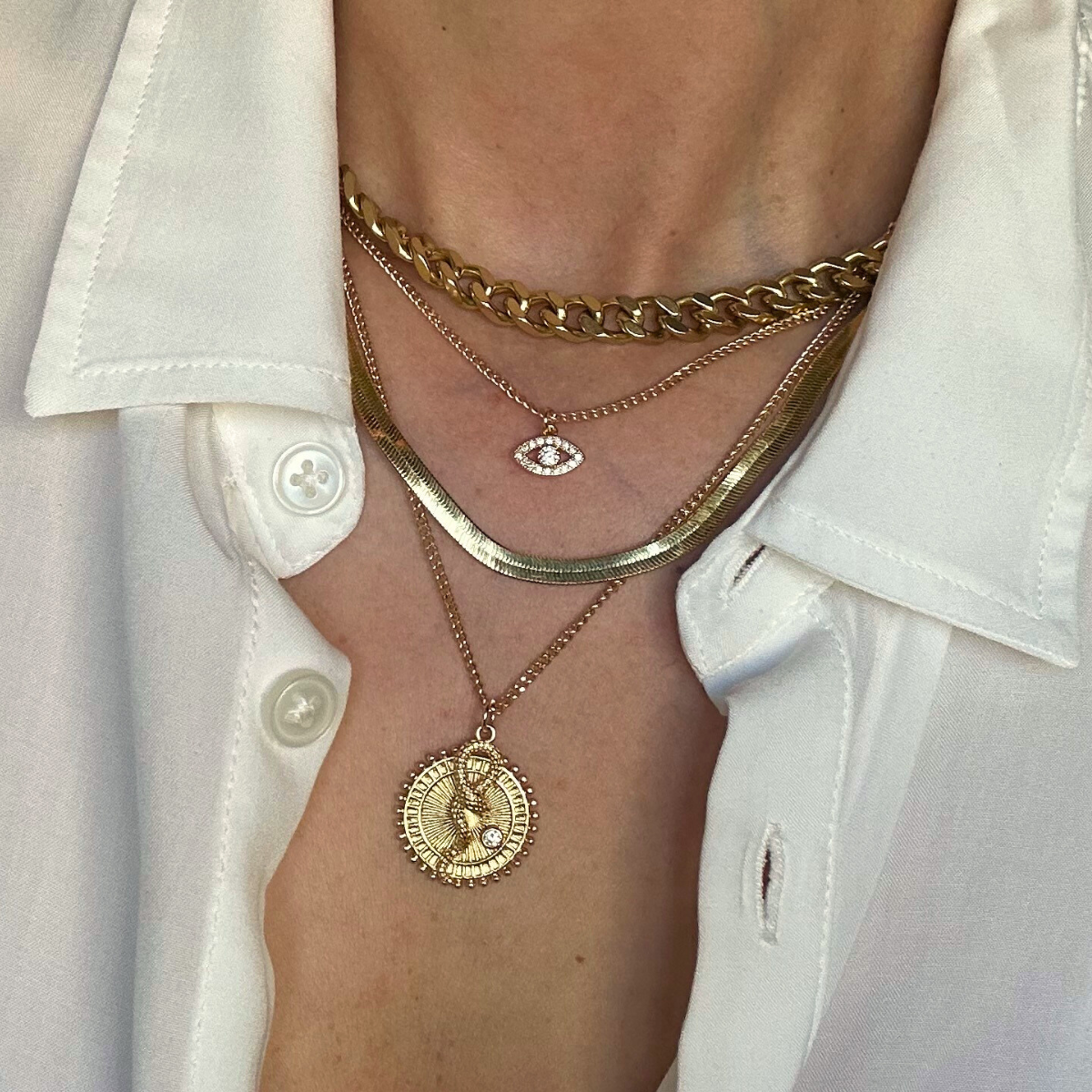 Close-up of a woman's neck and chest area showcasing multiple layered gold necklaces, including a chunky chain, a delicate chain with an eye-shaped pendant, a curved metallic collar, and a longer pendant with a circular design, worn over a white button-up shirt with an open collar.