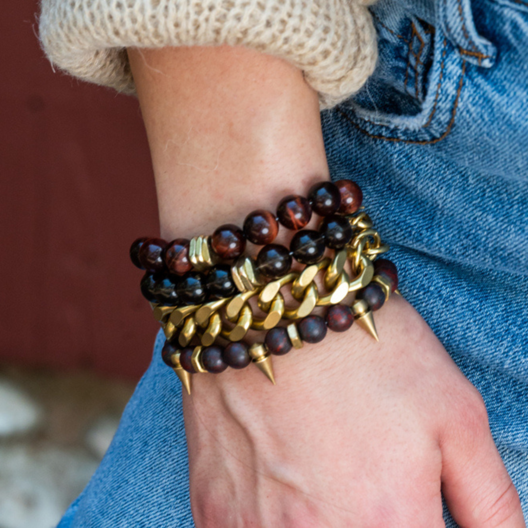 Close-up of a person's wrist wearing multiple bracelets, including brown beaded, gold chain, and gold spike accessories, with denim jeans and a beige knitted sweater.