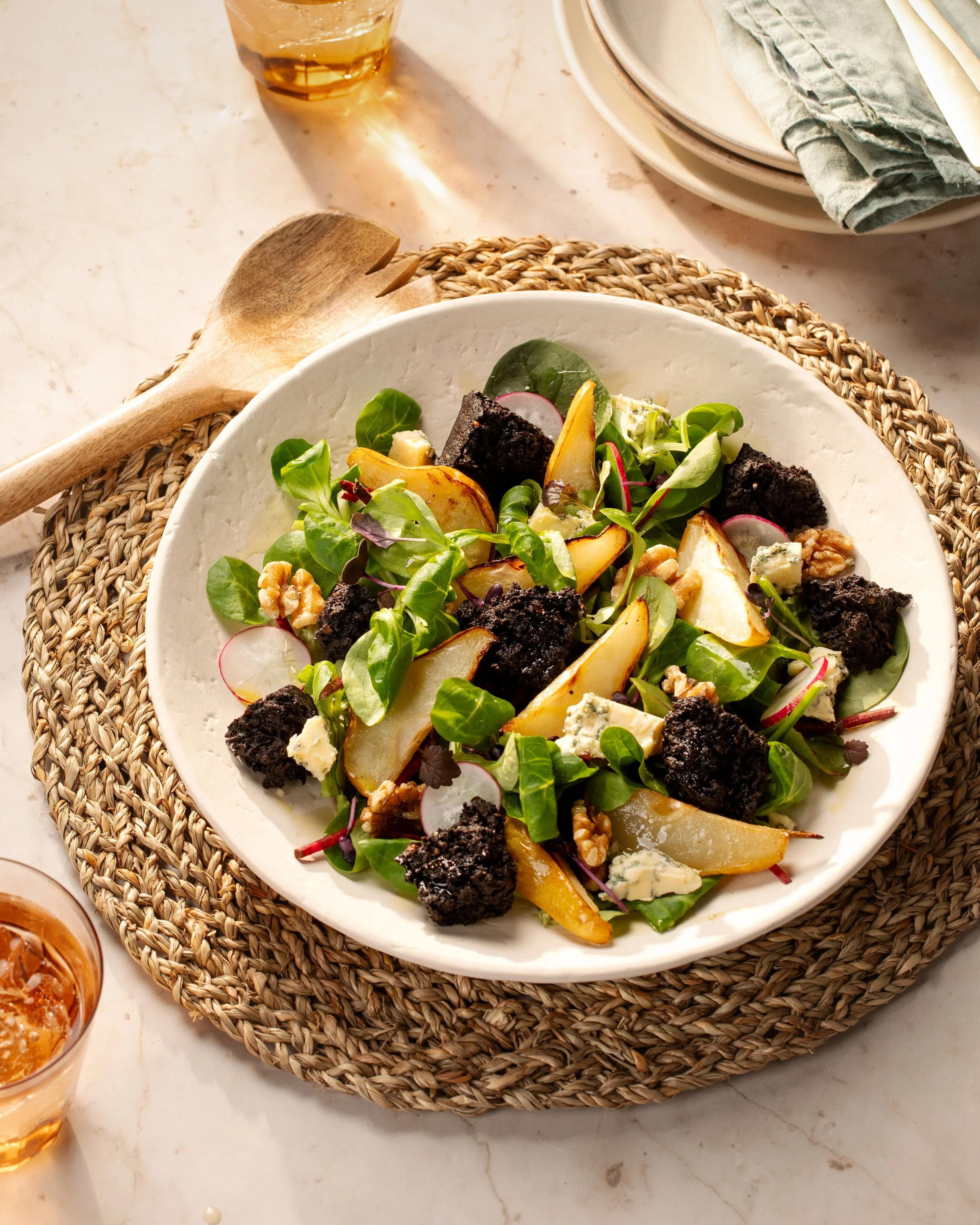 A bowl of mixed salad with greens, sliced pears, blue cheese crumbles, walnuts, and croutons on a woven placemat, with a wooden salad fork and glasses of drink nearby.