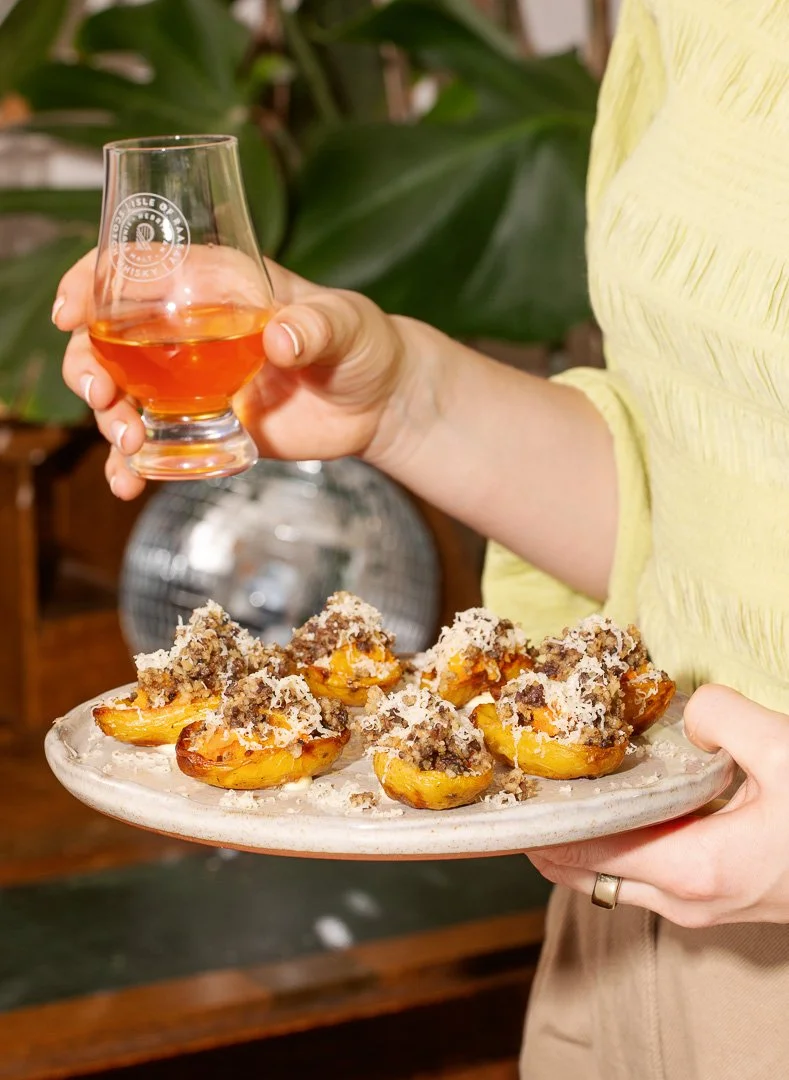 Person holding a plate of stuffed peaches topped with crumble and powdered sugar, and a glass of amber-colored drink.