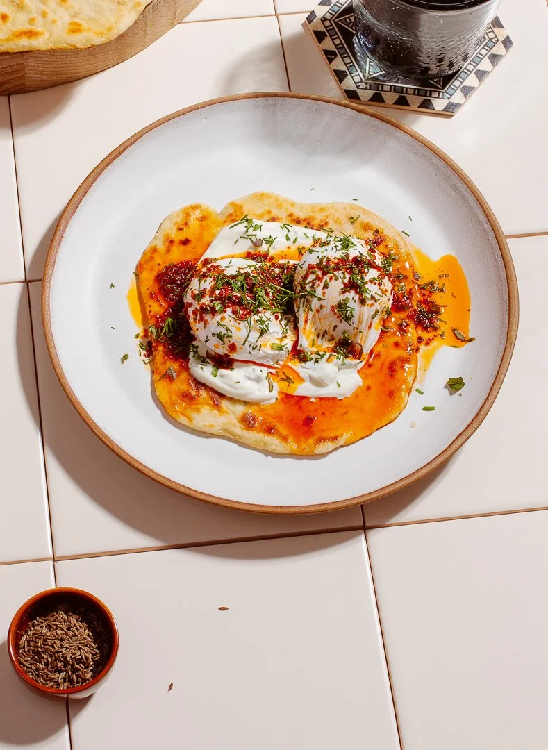 Poached eggs on flatbread with tomato sauce, garnished with herbs, served with a glass of soda on a tiled table.