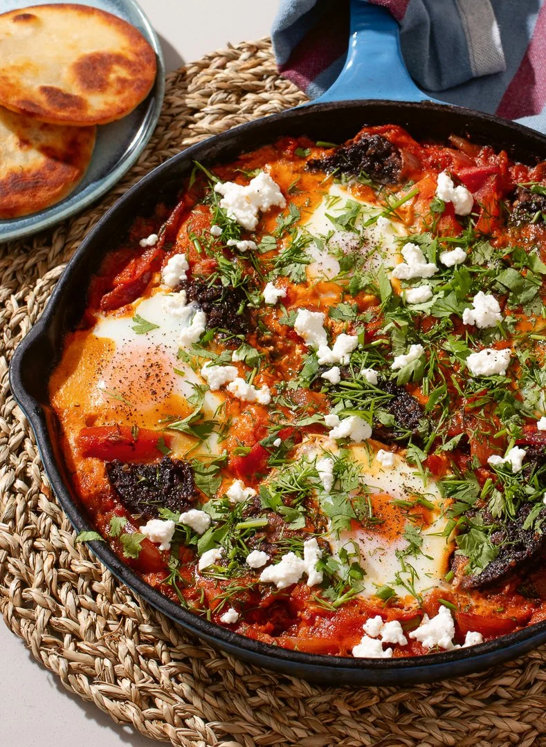 Baked eggs in tomato sauce with herbs, feta cheese, and black olives in a cast-iron skillet, with toasted bread on a plate nearby.