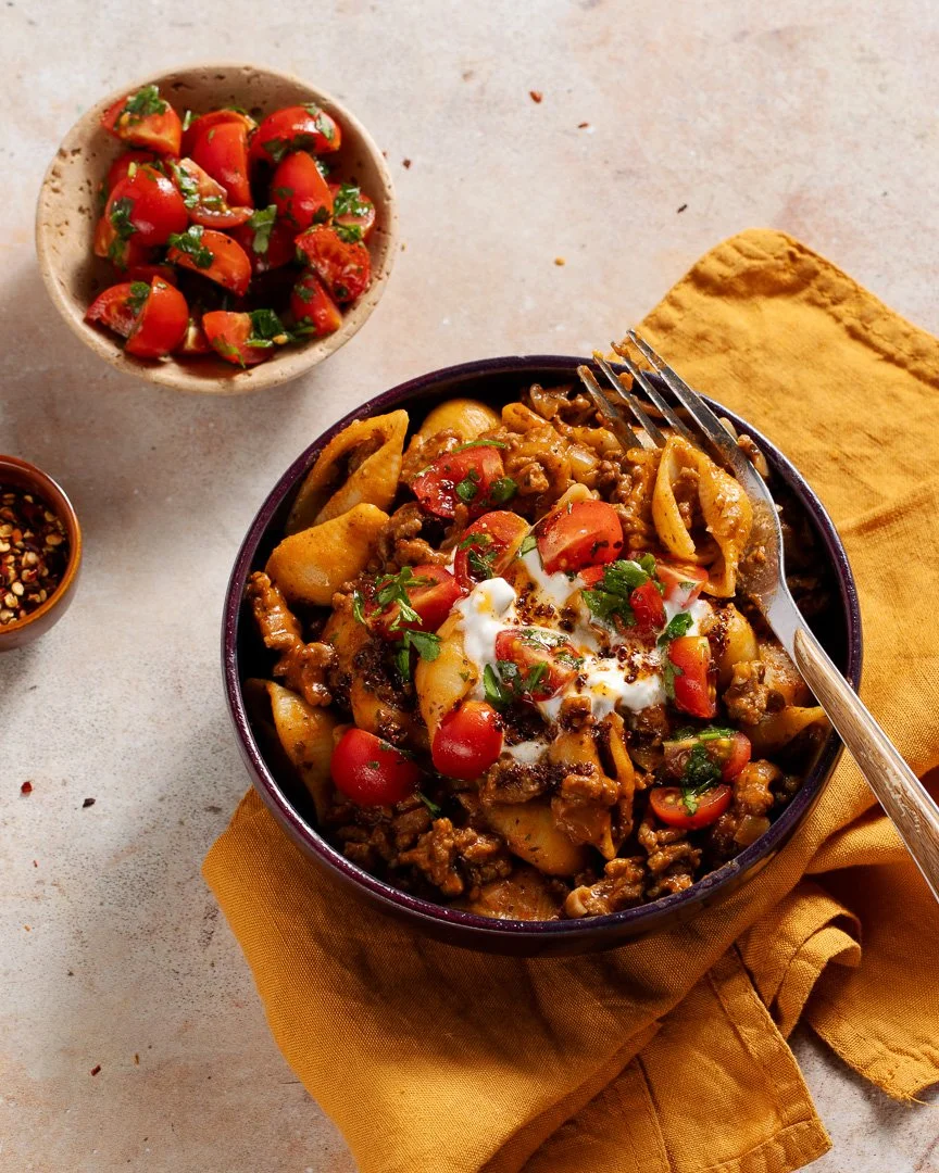 A bowl of taco meat with potatoes, cherry tomatoes, and herbs, topped with sour cream and chopped green onions, with a fork resting inside. A side dish of chopped tomato salad and a small bowl of red pepper flakes are on the table.
