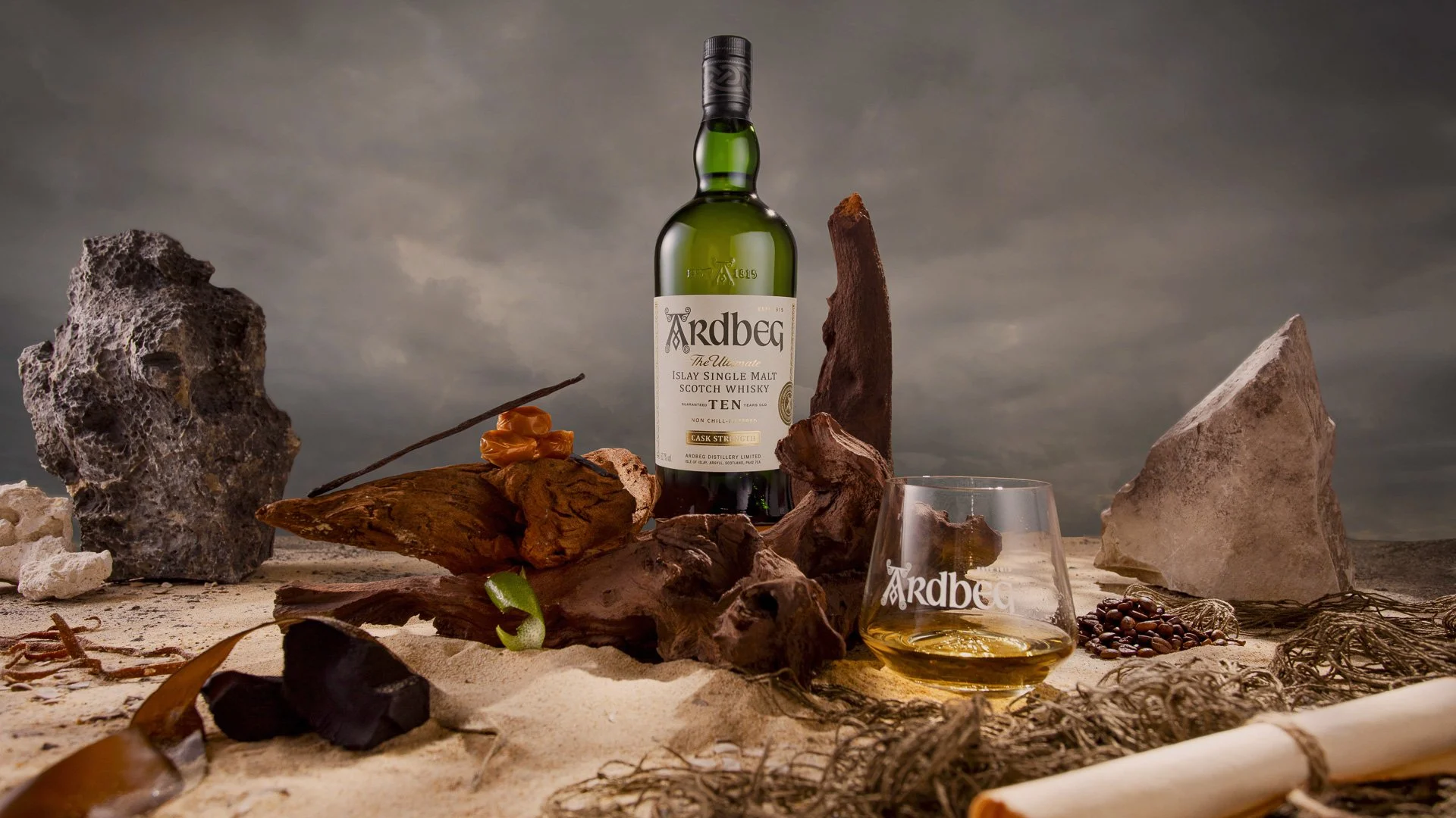 A bottle of Ardbeg scotch whiskey, a glass of whiskey, and various rocks, driftwood, and natural objects on sandy terrain under a cloudy sky.