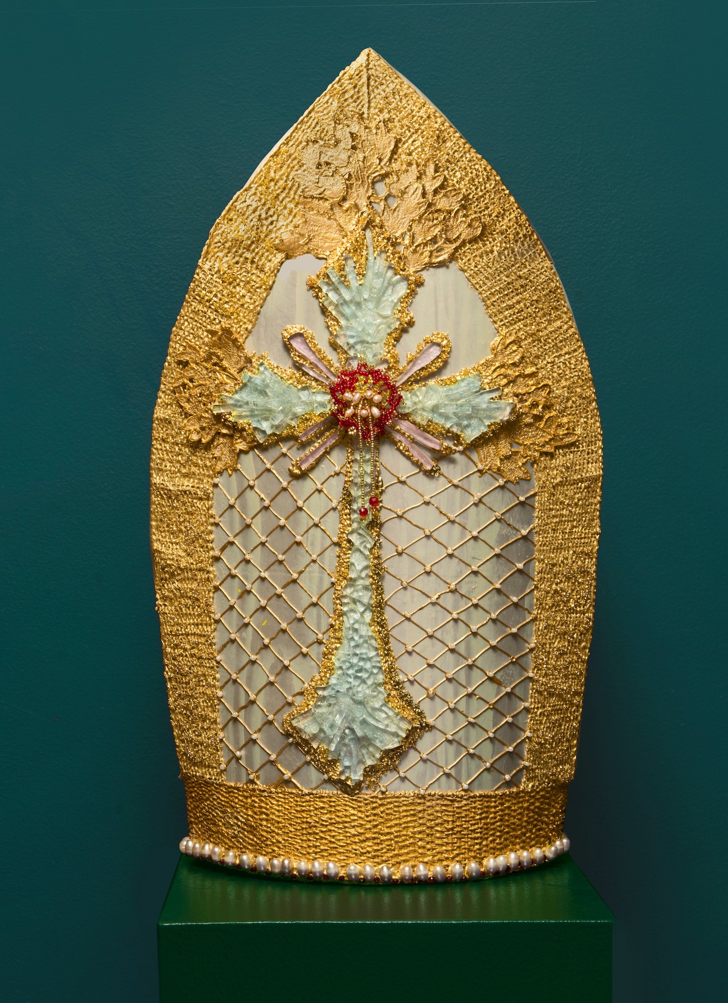 Front of a gold and silver mitre adorned with a cross of broken glass and beads.
