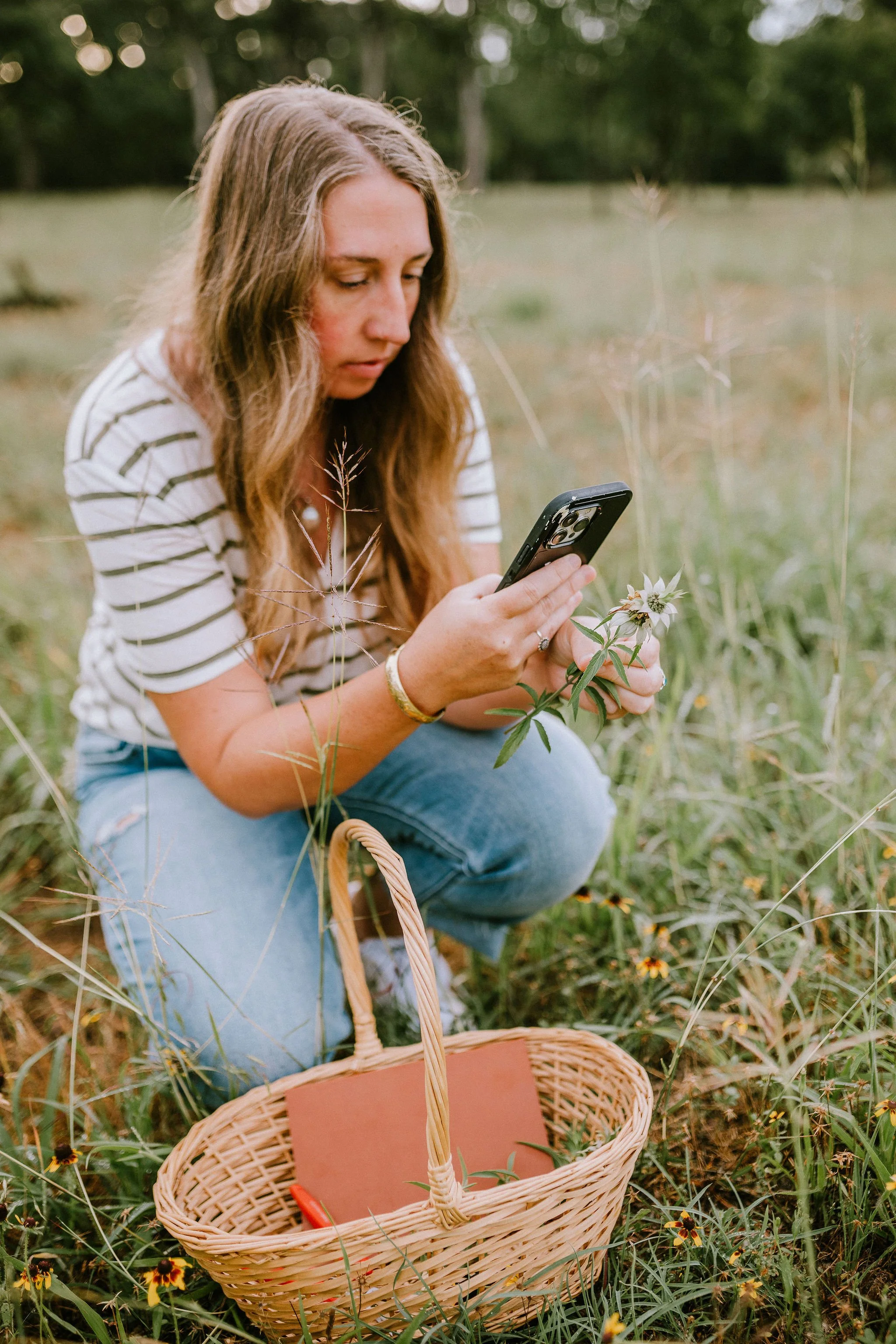 A woman with long wavy hair, wearing a white and black striped shirt and blue jeans, kneels in a grassy field while looking at her smartphone. There is a wicker basket on the ground nearby with a closed orange notebook inside and surrounded by wildflowers and grass.