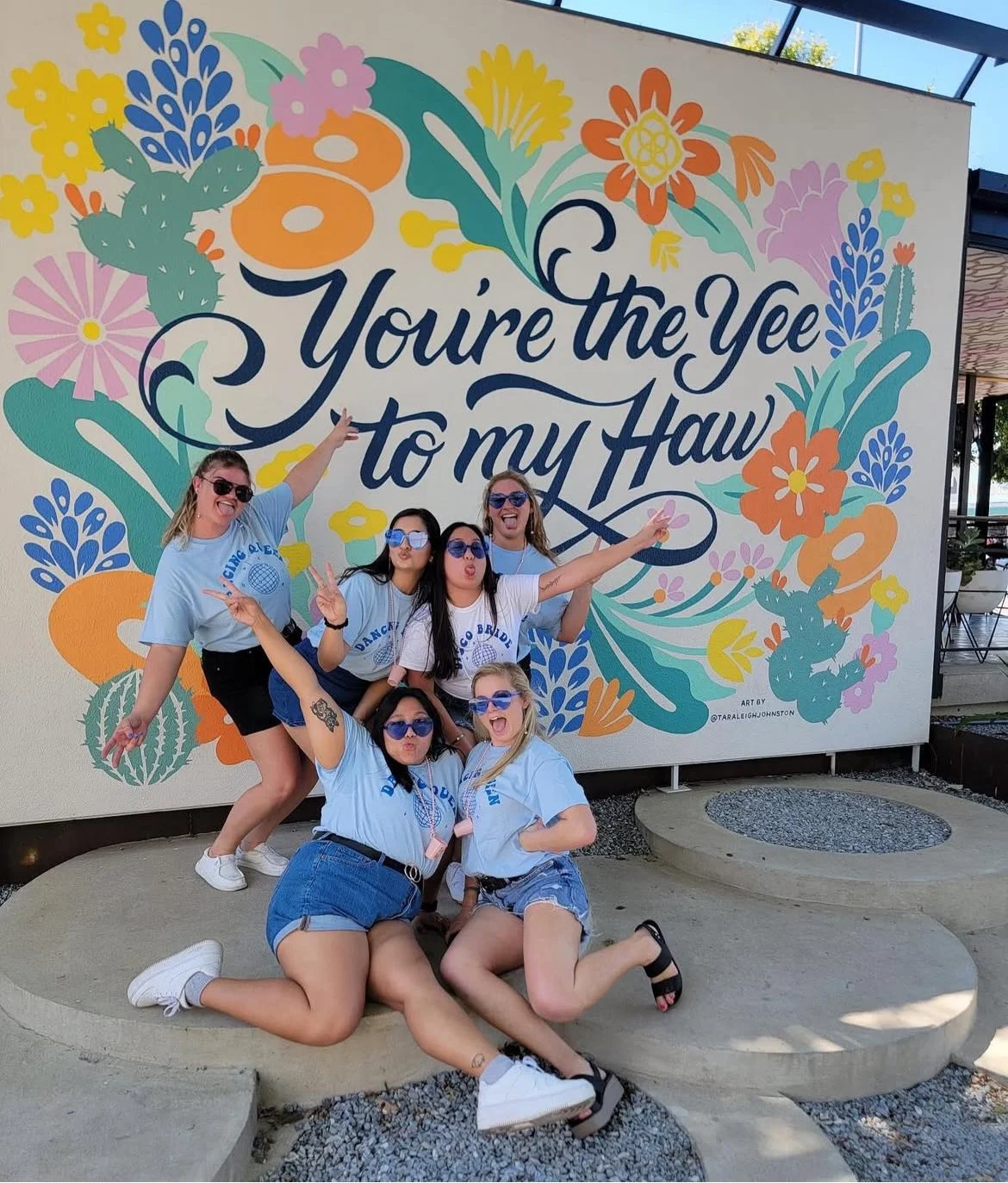 A group of six young women posing happily in front of a colorful mural that reads 'You're the Yee to my Haw,' with floral and cactus illustrations. They are wearing matching light blue T-shirts with 'DANCO BRIDE' and a globe logo, and all are wearing sunglasses, making playful expressions and peace signs.