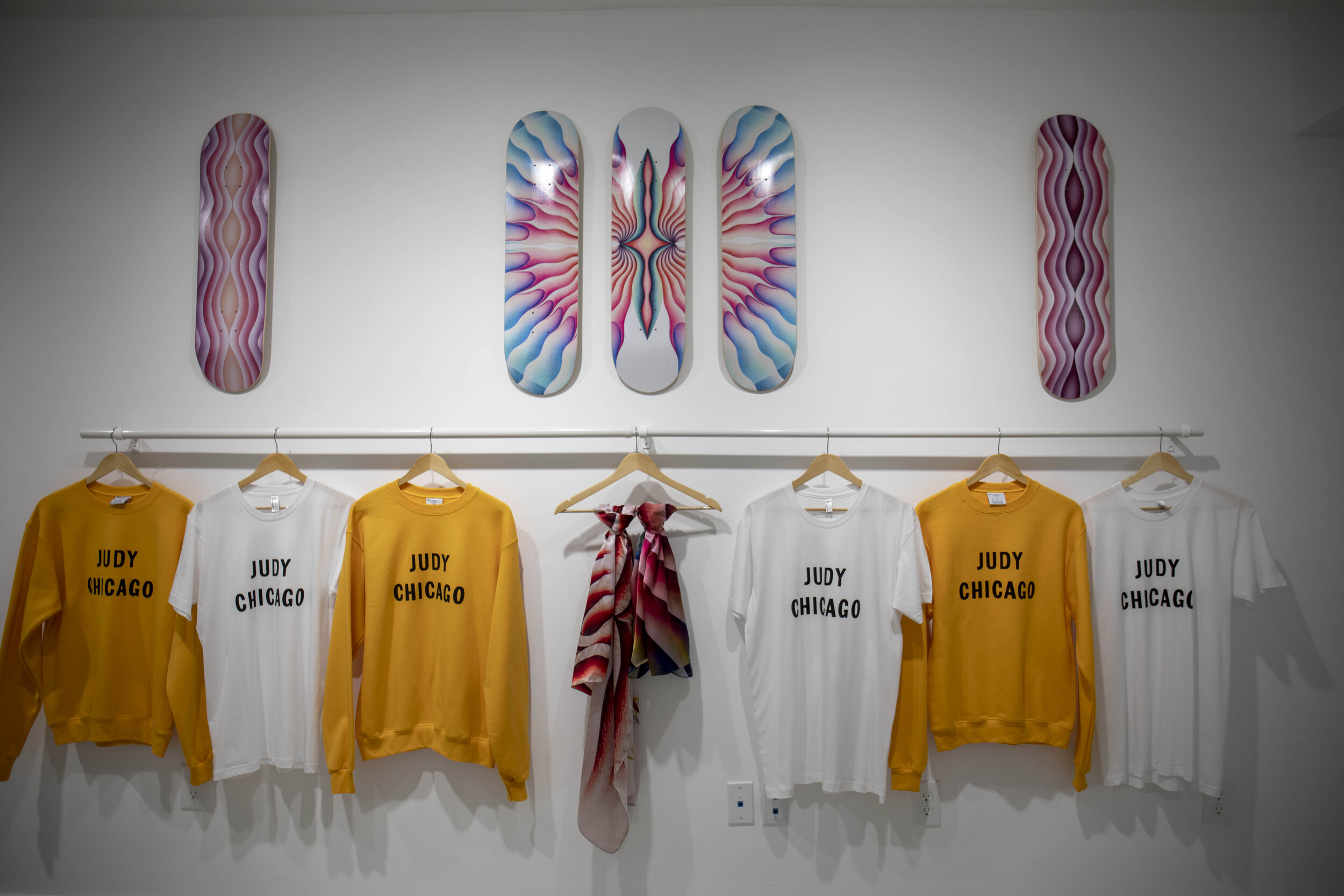  Judy Chicago sweatshirts align the wall in the shop portion of Through the Flower Art Space.  Above the sweatshirts are skate boards designed by Judy Chicago. From left to right, The Light Ripple skate board, Butterfly Ripple skateboard, and the Dar