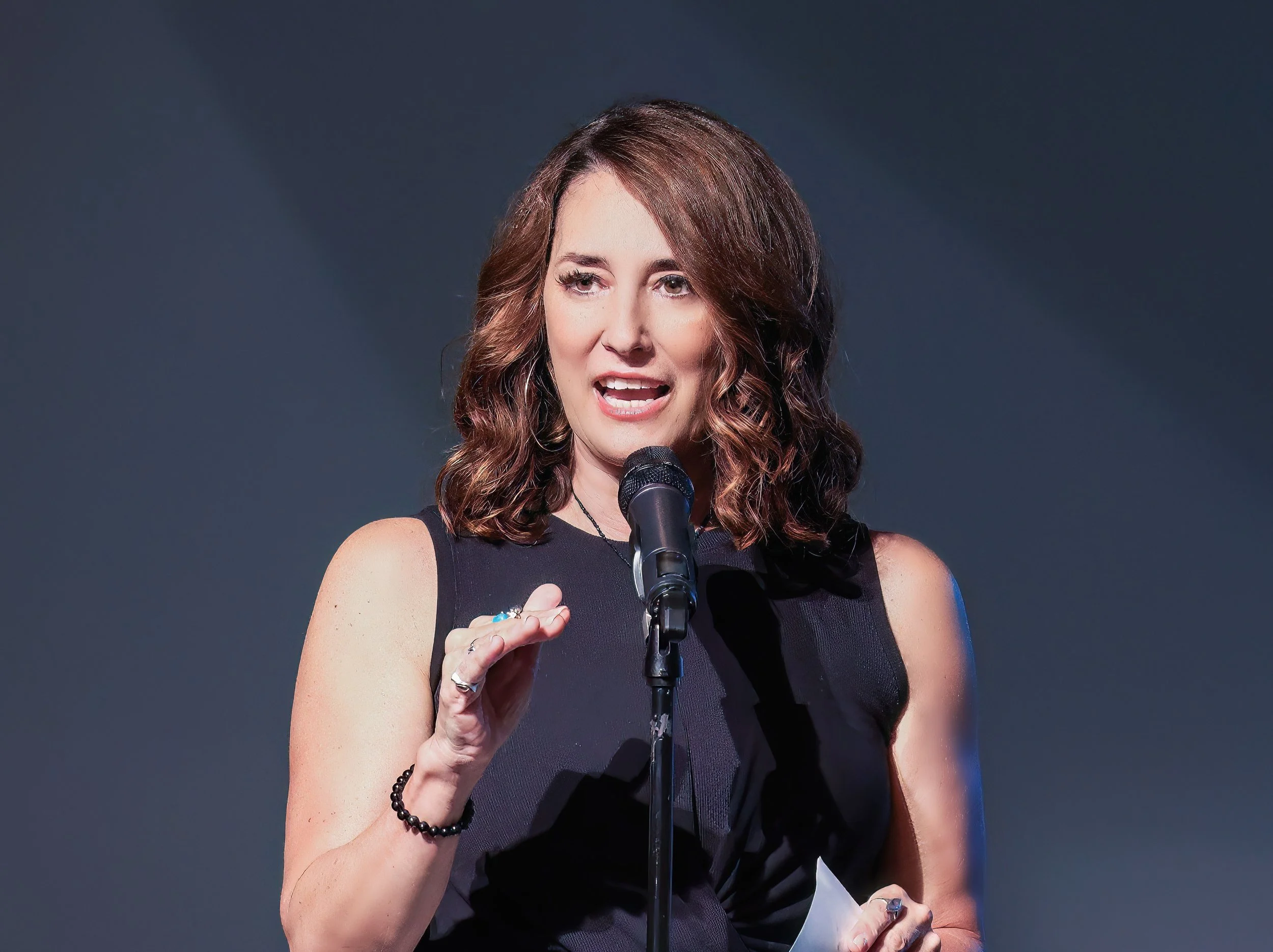 A woman with shoulder-length, wavy brown hair speaking into a microphone while holding a piece of paper, wearing a sleeveless black top, against a dark background.