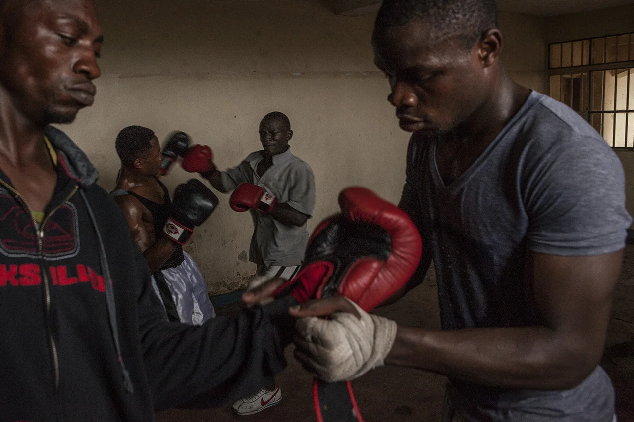 Fabrice and Claude, two boxing students, help each other to put on their gloves while Kibomango spars with another student in the background. Goma, North Kivu, DRC