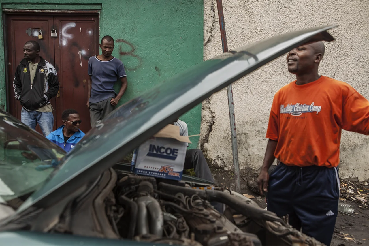 Freelance mechanics work on a car in the street at Kibabi in Goma. Kibomango is forced to work as a mechanic and a security guard because, despite being the champion of the DRC, boxing earns him no income. The mechanics cannot afford to rent a garage so are forced to work out in the street, whatever the weather. Goma, North Kivu, DRC