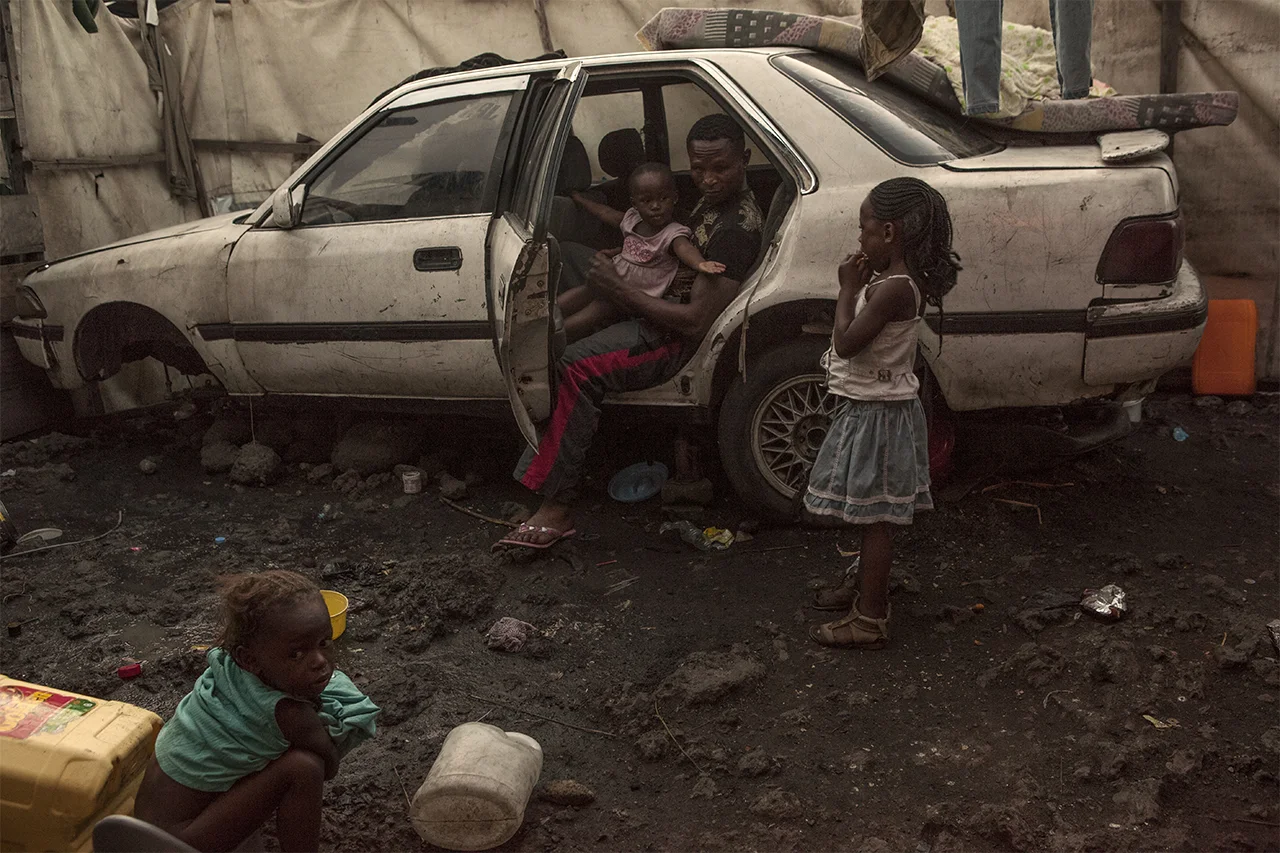 Kibomango playing with his children while sat in his broken-down car in front of his home. Goma, North Kivu, DRC