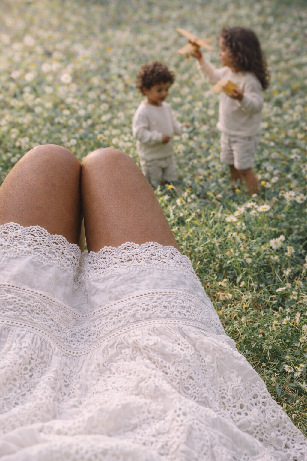 Woman, mother laying down in wildflowers field in a white lace cotton dress, watching her two children playing