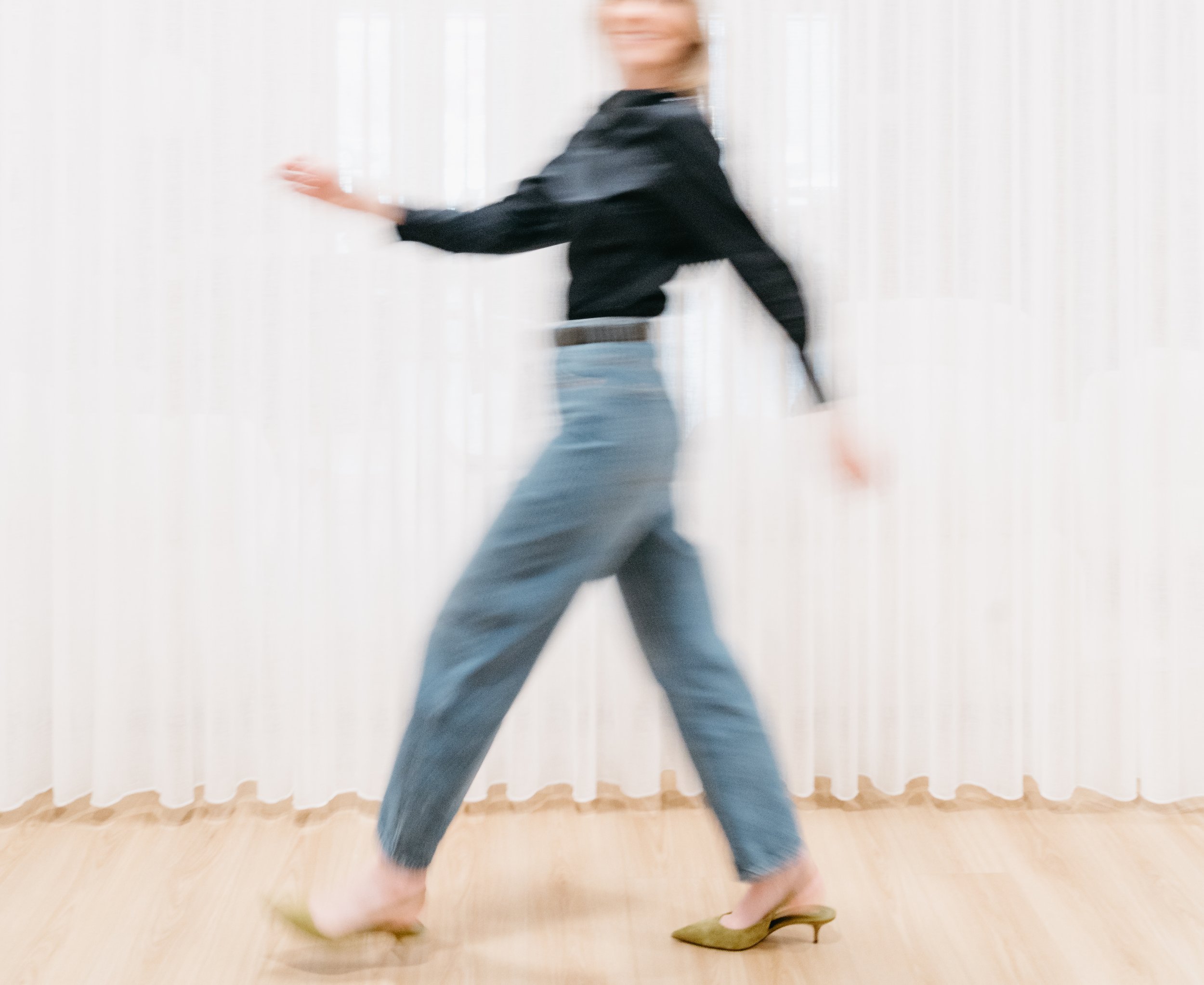 A blurred image of a person dancing indoors with white curtains in the background. The person is wearing a black top, blue jeans, and beige shoes.