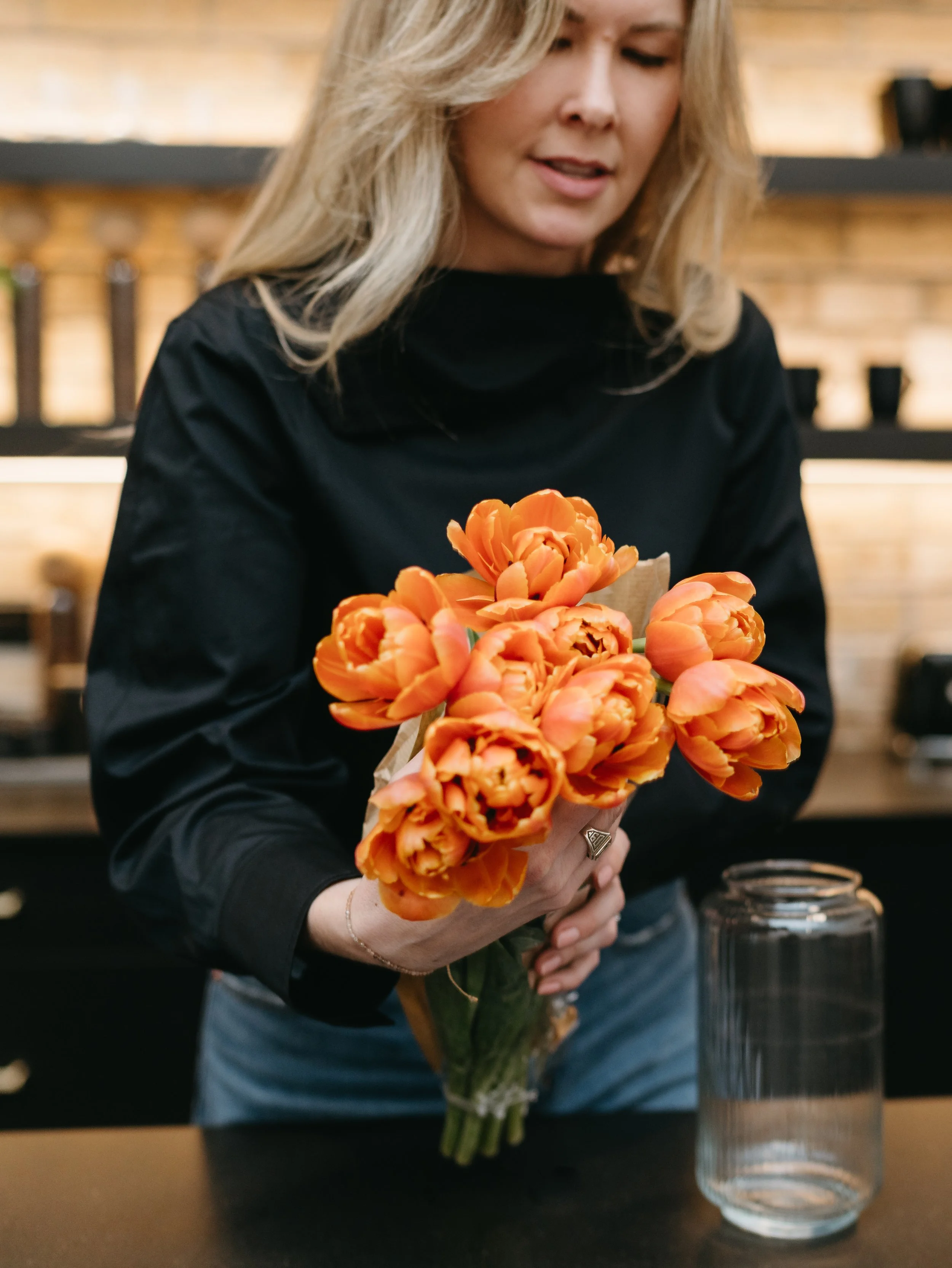 A woman with blonde hair in a black top holding a bouquet of orange tulips in a glass jar in a cozy indoor setting.