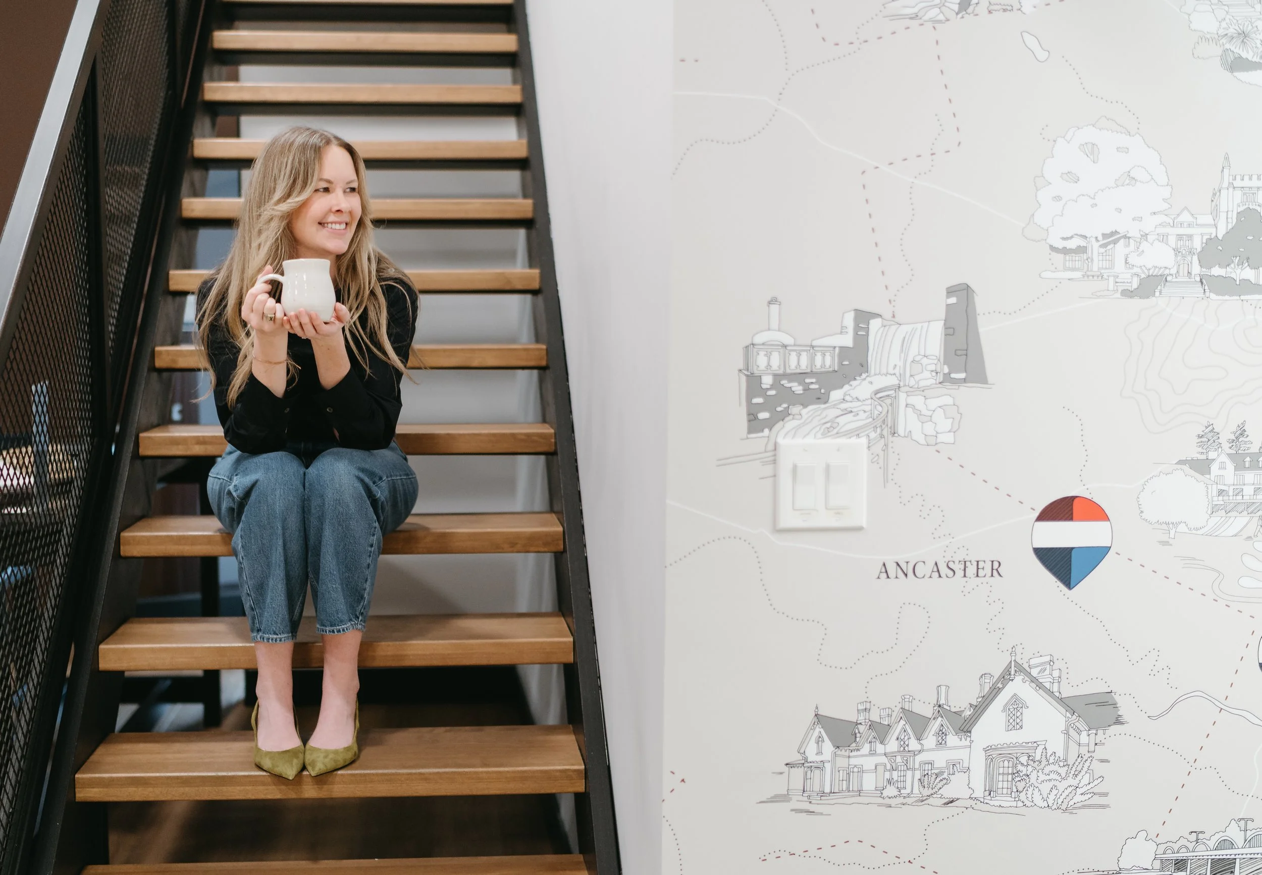 A woman sitting on wooden stairs, holding a white mug, smiling, with a wall map of Lancaster behind her.