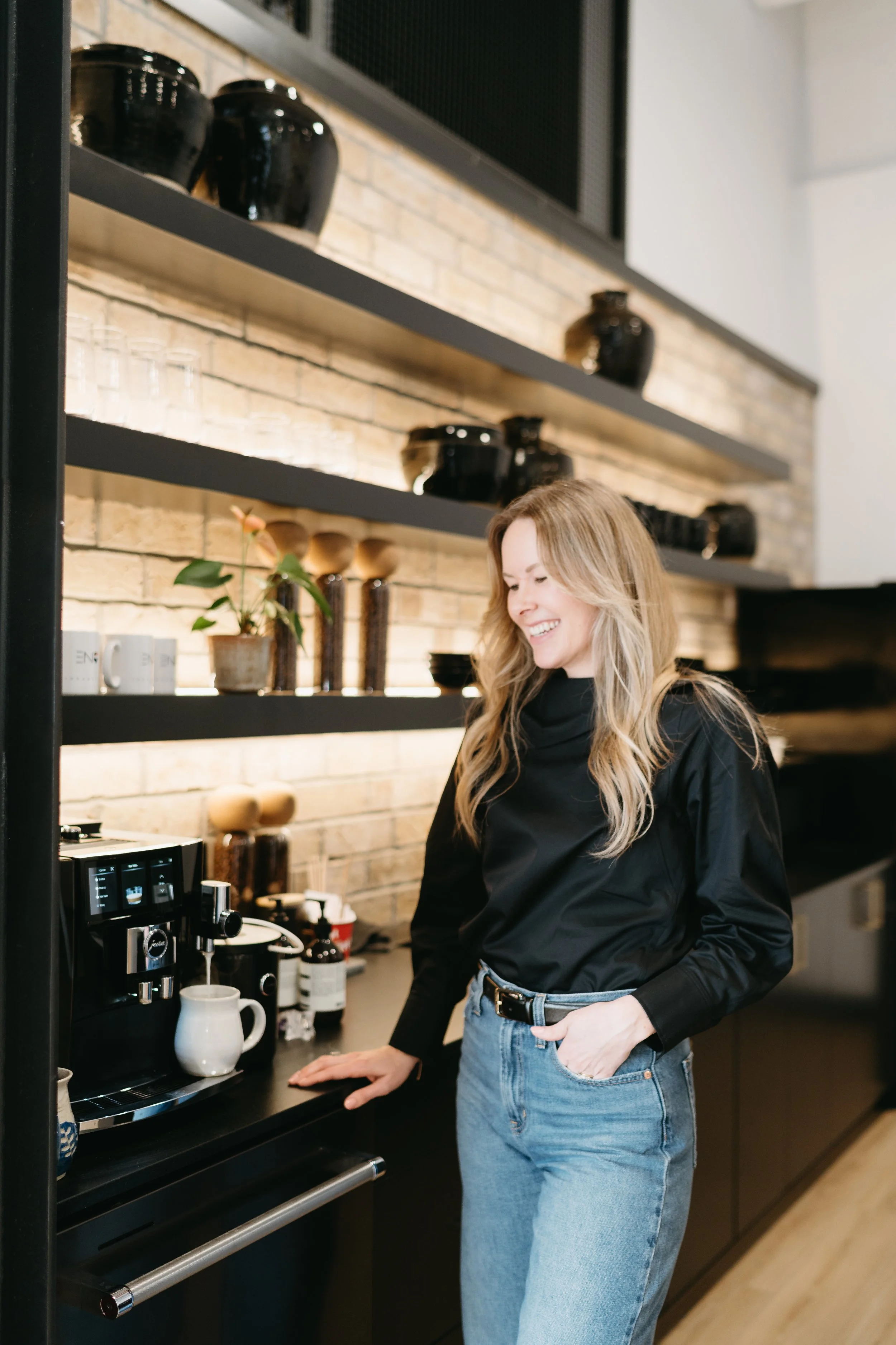 A young woman with long blonde hair wearing a black top and blue jeans, smiling, standing in a modern kitchen next to a coffee machine and a brick wall with black shelves holding black pots and jars.