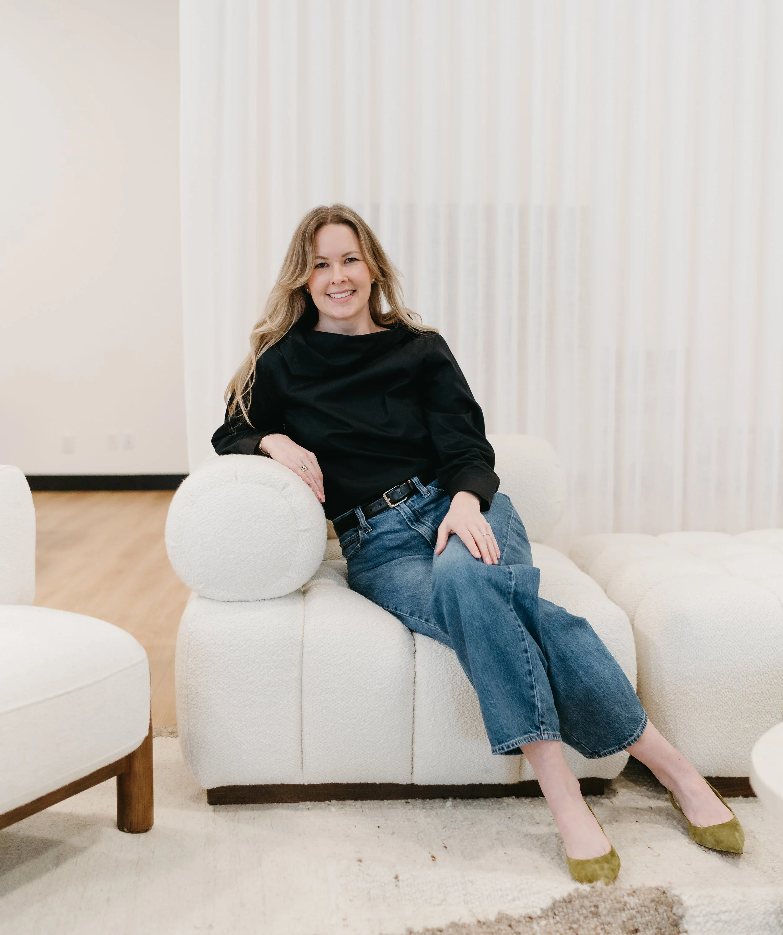 A woman sitting on a white sofa in a modern, minimalistic room. She has long, wavy blonde hair and is wearing a black top, blue jeans, and green high heels. She is smiling at the camera with one arm resting on a round pillow.