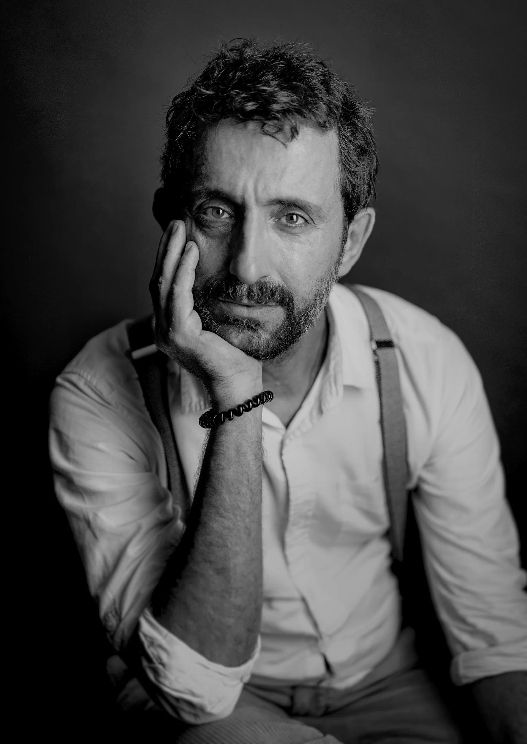 Black and white portrait of a man with a beard and curly hair, resting his face on his hand, wearing a light-colored shirt with rolled-up sleeves and suspenders.